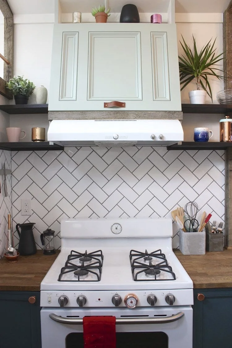 A kitchen cooking area features a white gas stove, a white herringbone tile backsplash, and a sage green range hood cover flanked by dark wood shelves.