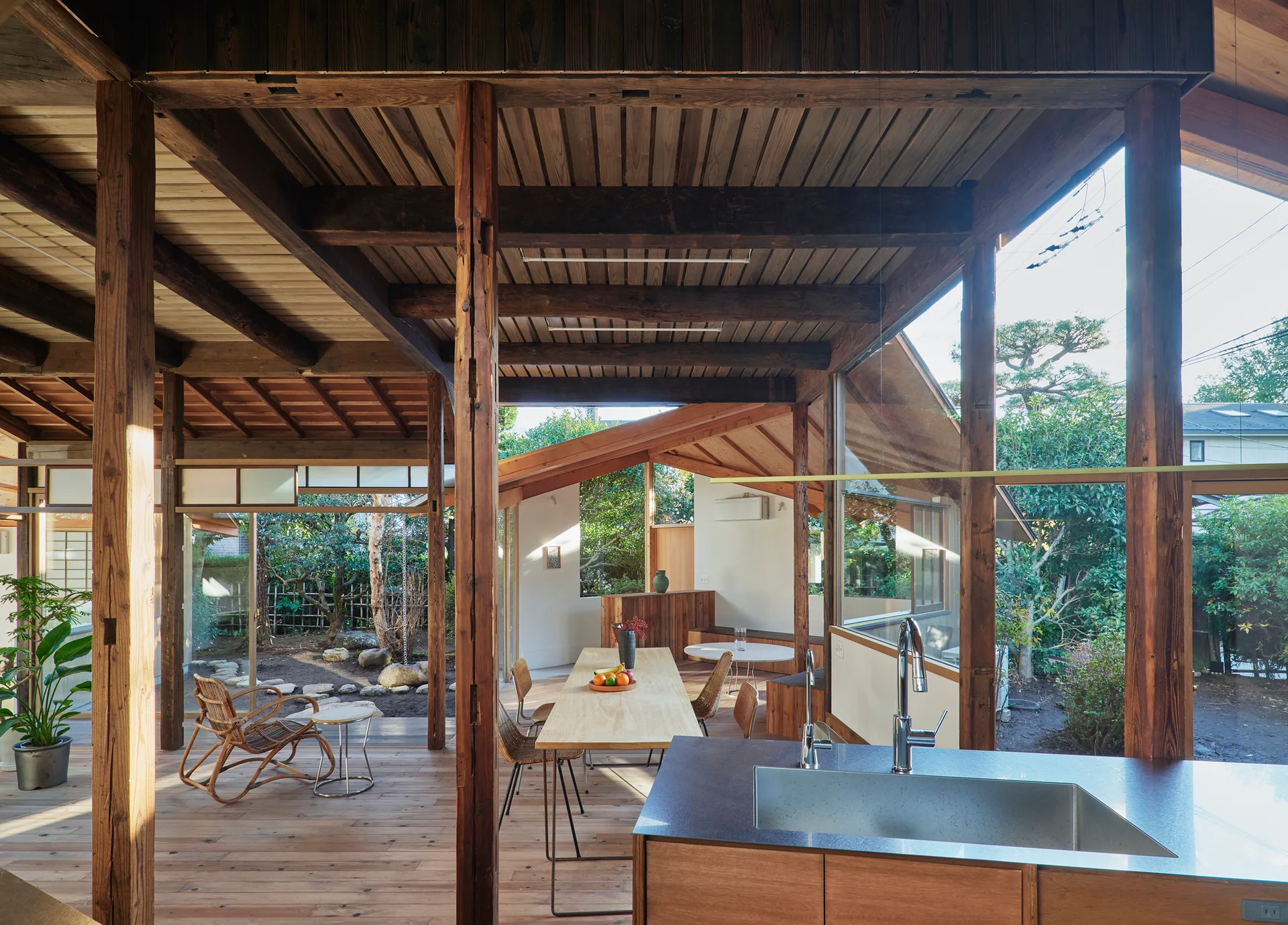 An open plan kitchen and dining room featuring exposed timber beams, a light wood dining table, and large glass windows overlooking a garden.