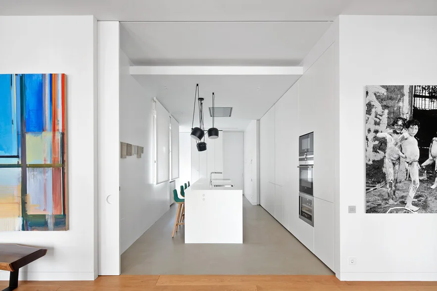 A minimalist white kitchen features a central white island with turquoise bar stools, integrated wall cabinets, and large artwork on the flanking walls.