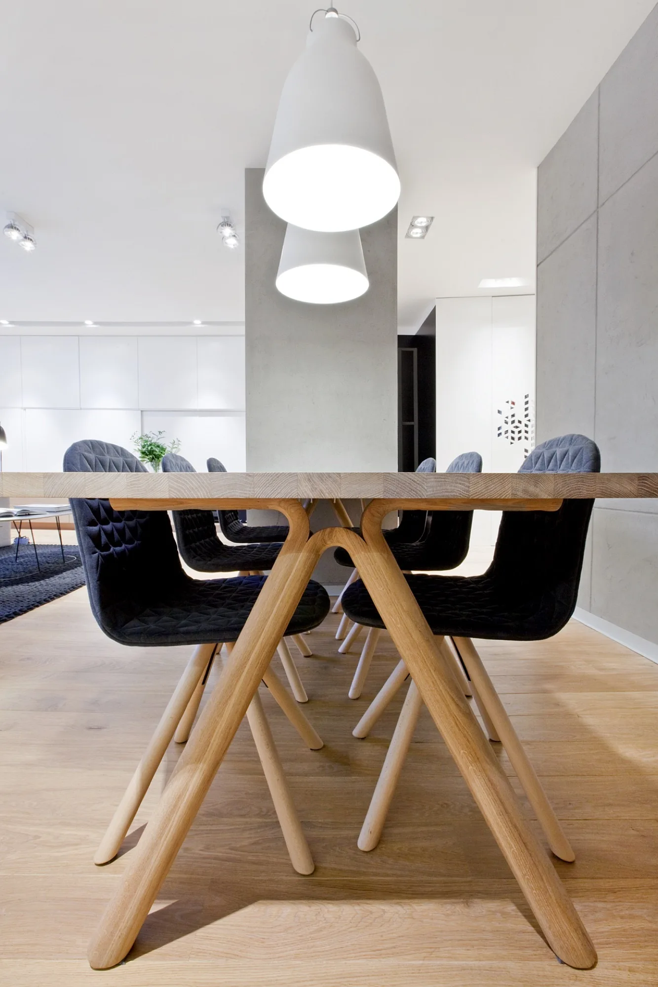 A wooden dining table with angled legs is surrounded by dark quilted chairs under two white pendant lights in a modern dining room.