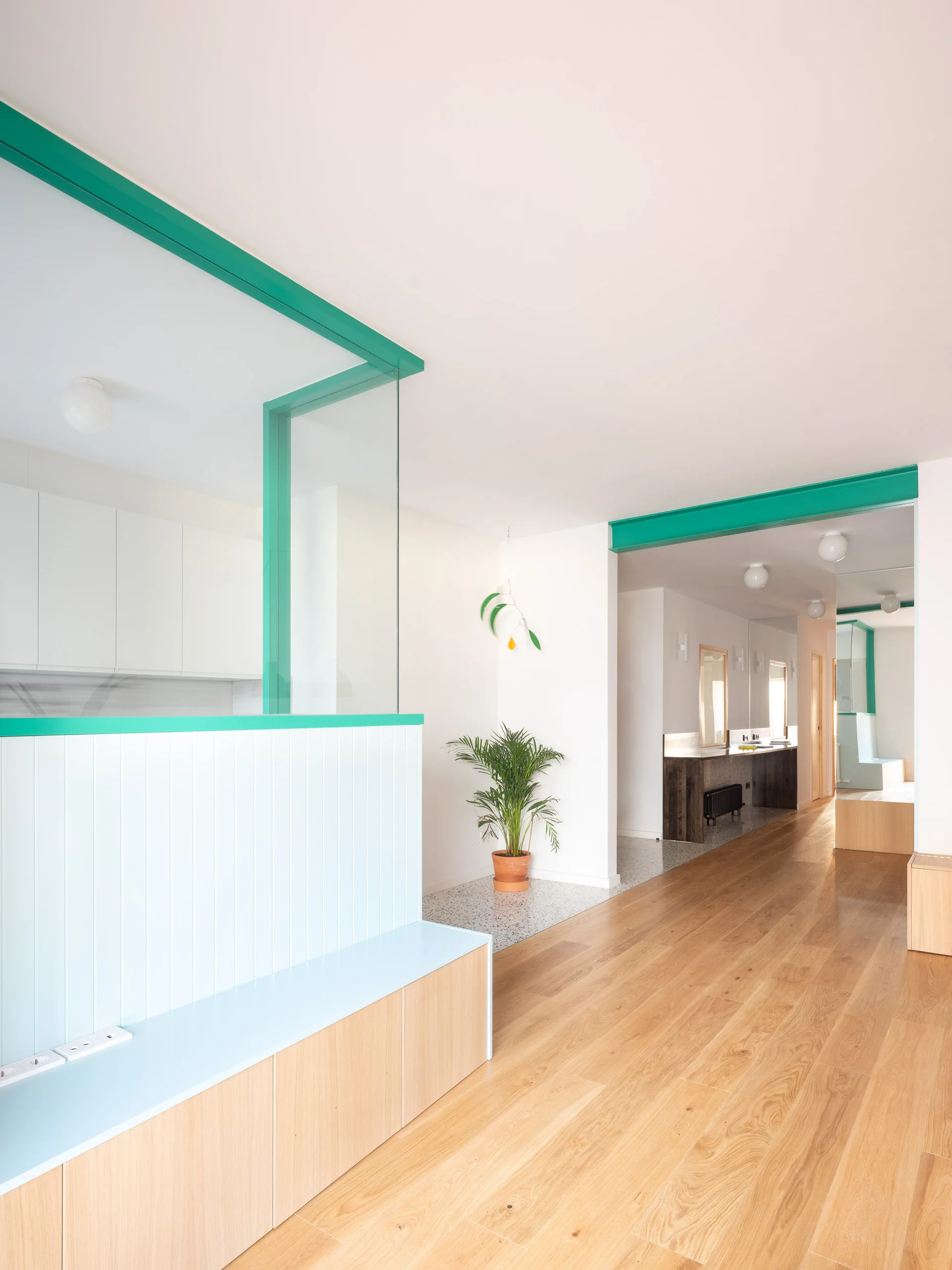 A modern residential hallway with wide plank wood floors, a light blue paneled divider, a teal-framed glass partition, and ceiling globe lights.