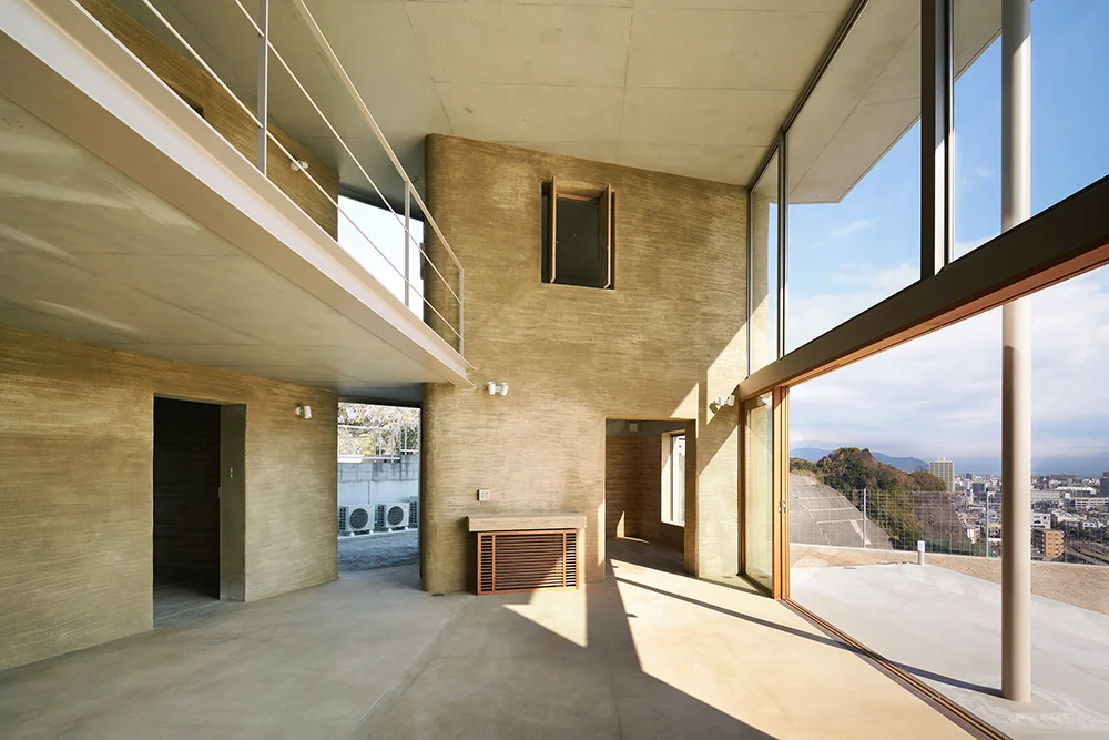 A minimalist interior features rough earth-textured walls, a smooth concrete floor and ceiling, a mezzanine level, and large glass windows.