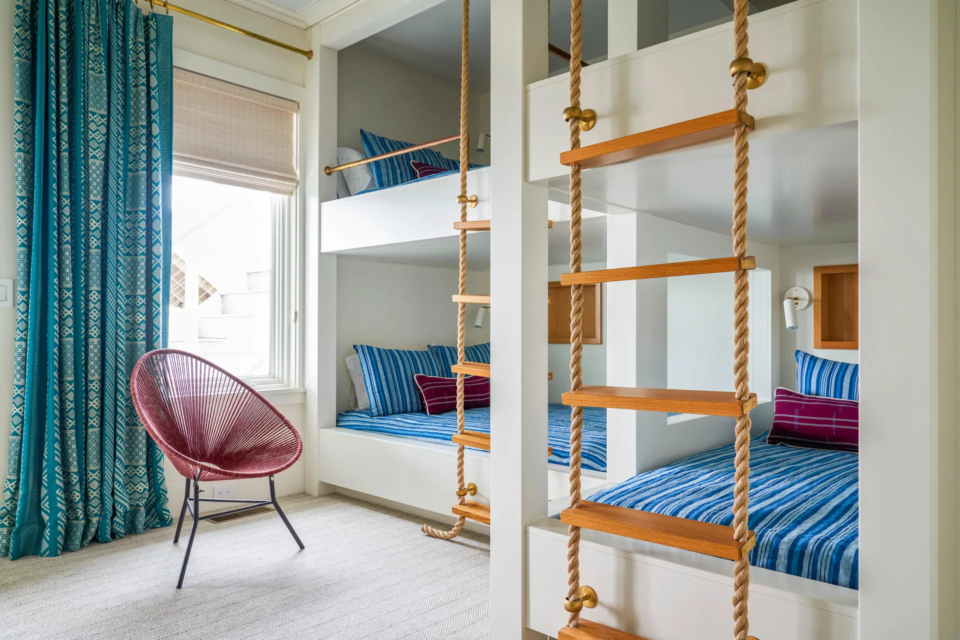 A kids' bedroom featuring custom white built-in bunk beds with thick rope ladders, blue striped bedding, and a red woven accent chair.