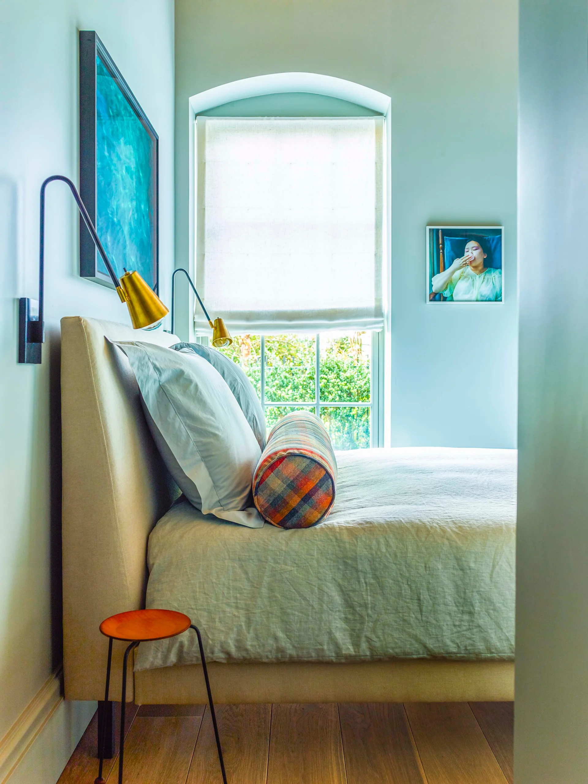 A light blue bedroom featuring an upholstered beige bed with a plaid bolster pillow, brass wall sconces, and an arched window with a roman shade.
