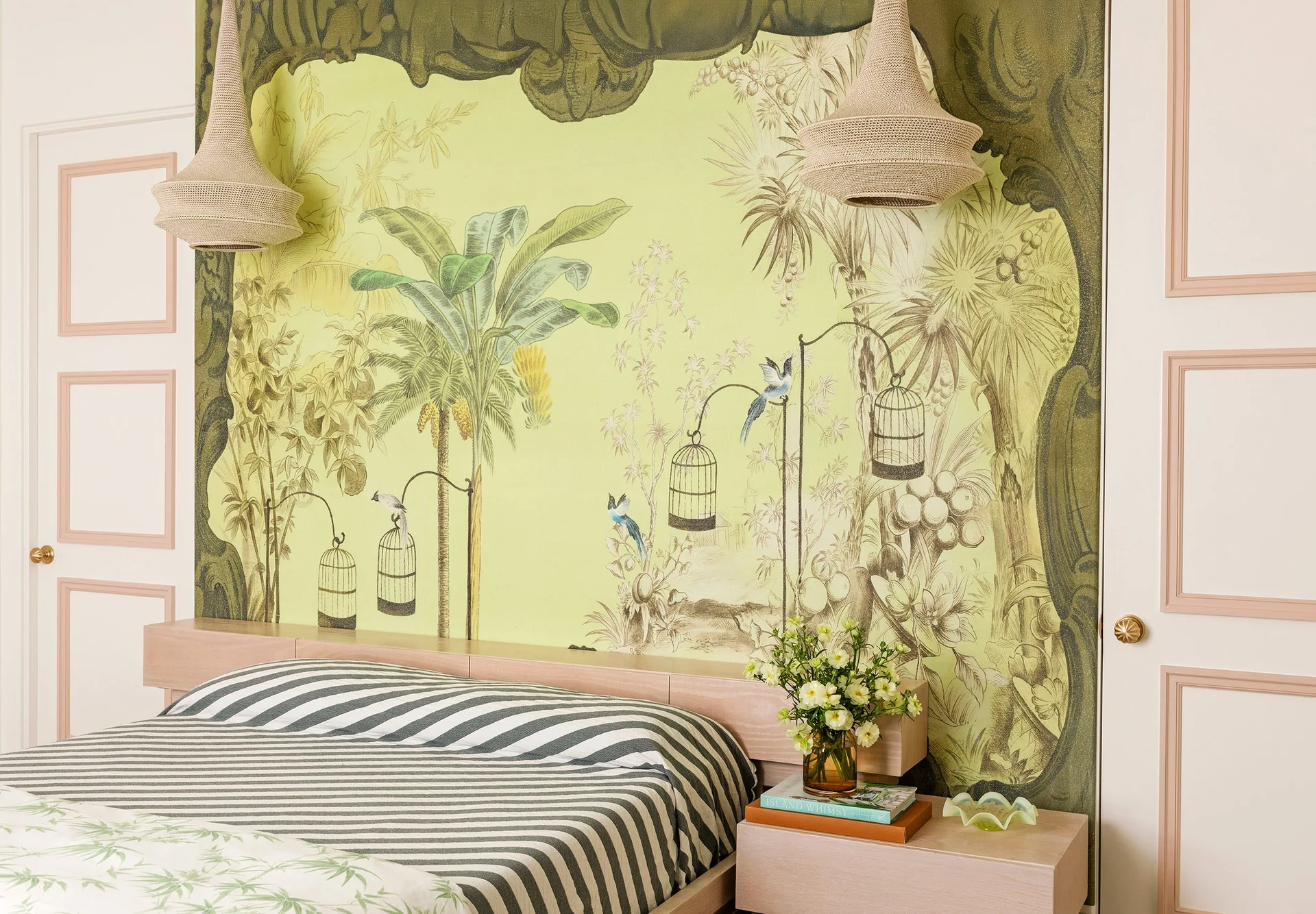 A bedroom featuring a large tropical green mural behind a low wooden bed with striped bedding and white flanking doors detailed with soft pink trim.