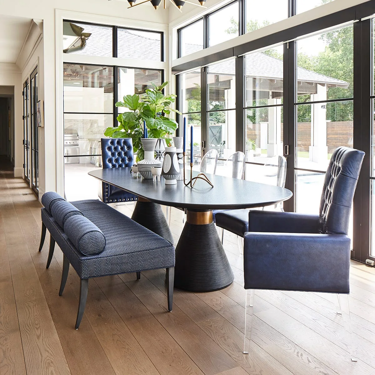 A modern dining room featuring a black oval table with a navy upholstered bench, tufted blue leather chairs, and large black-framed windows.