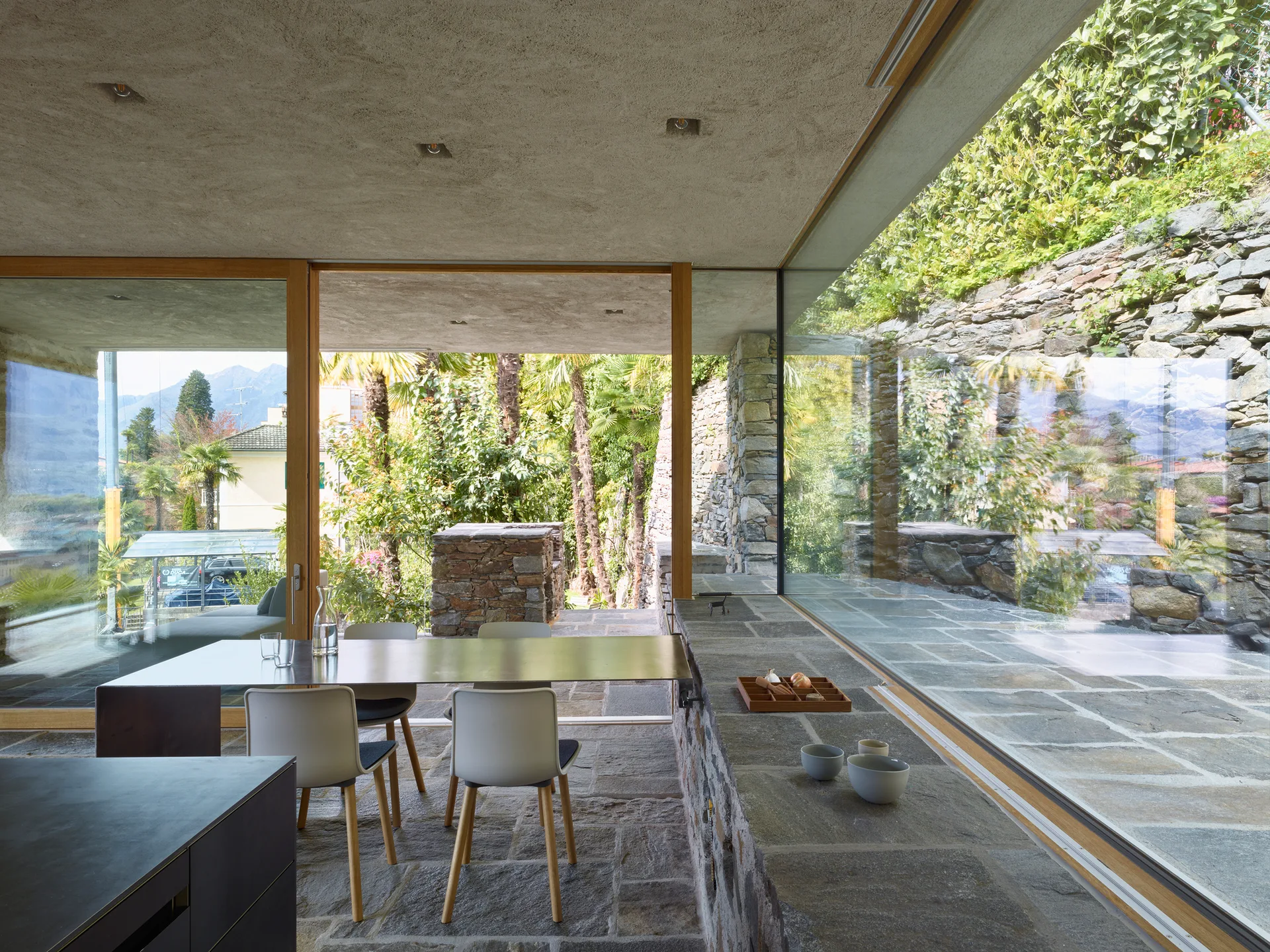 A minimalist dining room with a metal table and white chairs features grey stone flooring and large glass walls overlooking a garden.