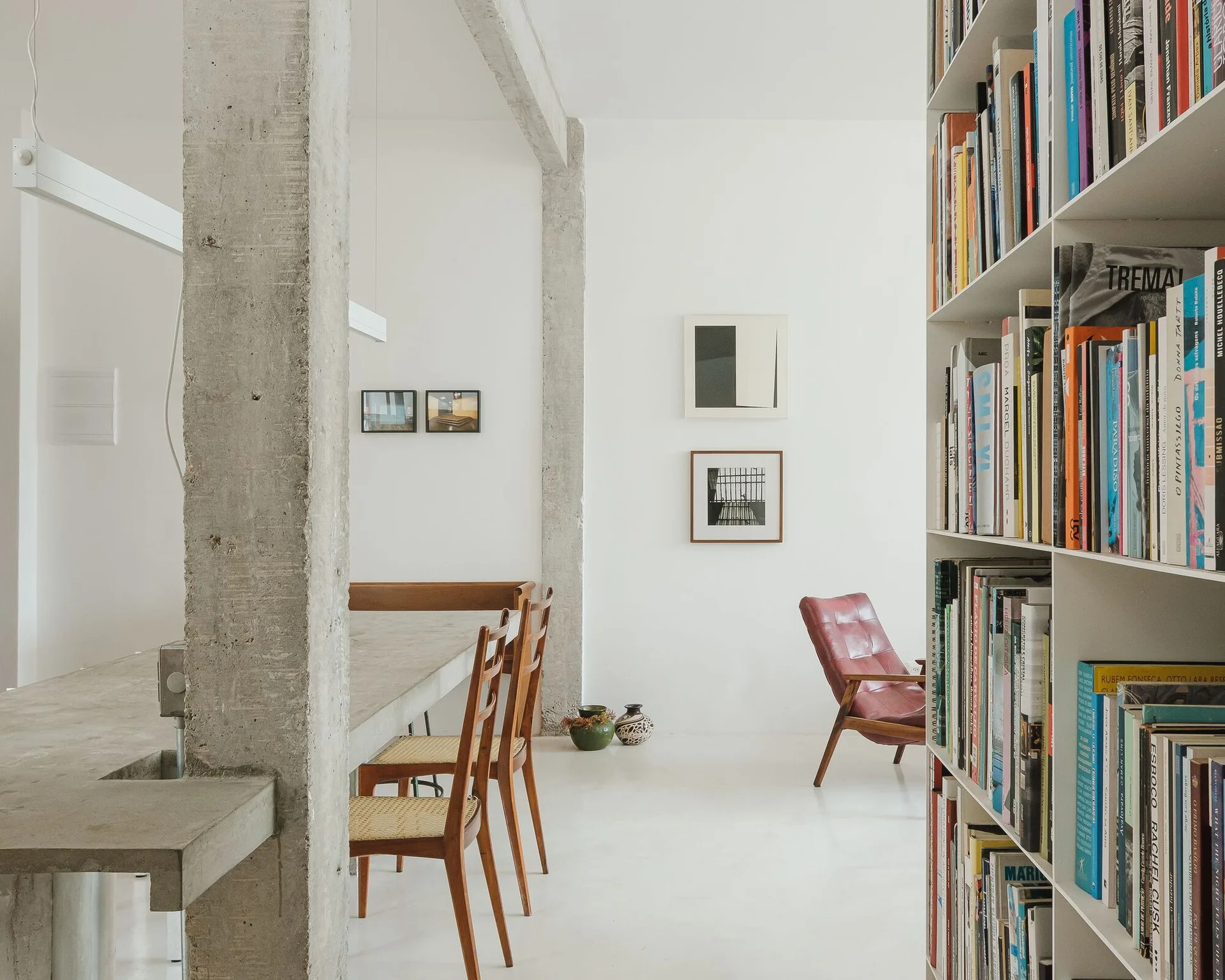 A minimalist dining area features an exposed concrete pillar and table, wooden chairs with cane seats, and a tall white bookshelf filled with books.