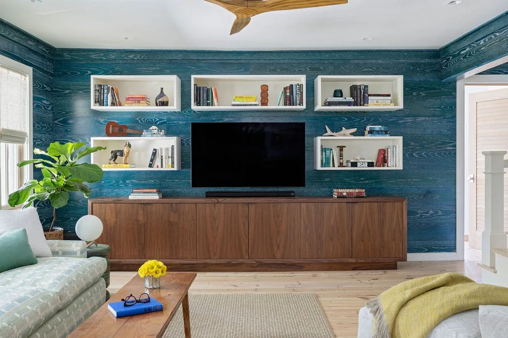 A living room features a blue wood-grain accent wall with white floating box shelves, a mounted TV, and a long wooden media console.