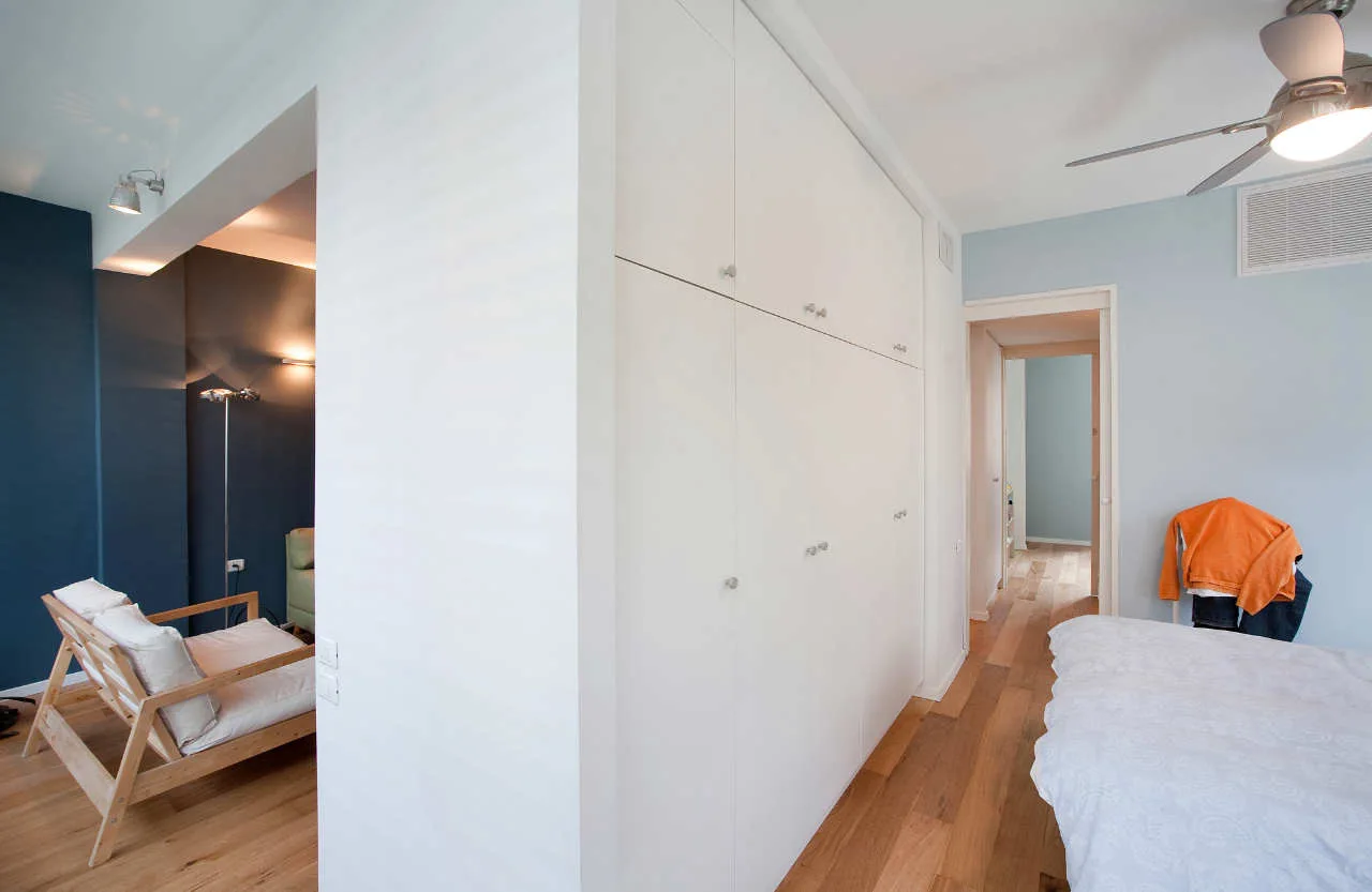A minimalist bedroom featuring a massive floor-to-ceiling white wardrobe, light wood flooring, and an adjoining room with deep blue walls.