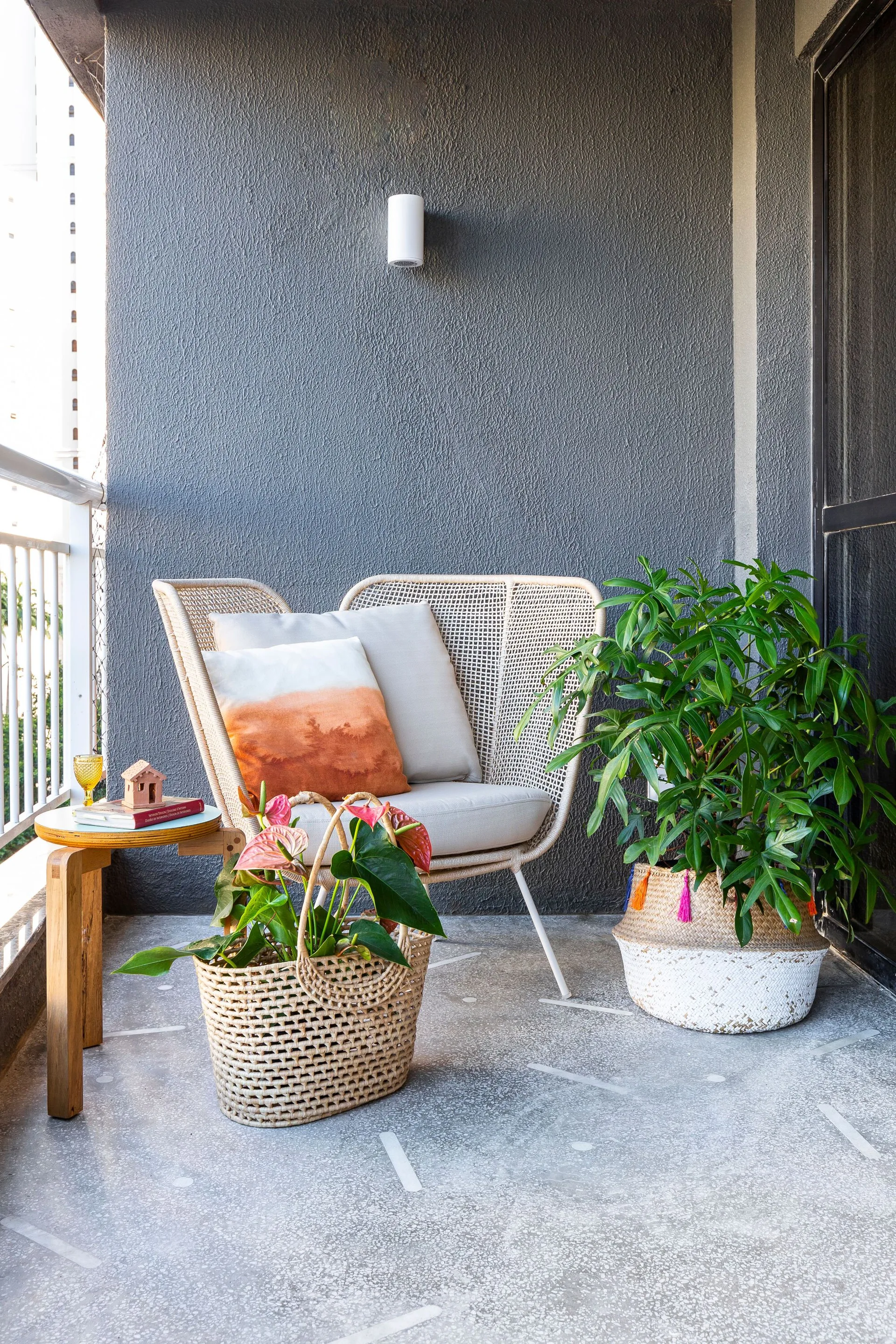 A cozy balcony corner features a wicker armchair with light-colored cushions next to green plants housed in woven baskets against a dark gray wall.