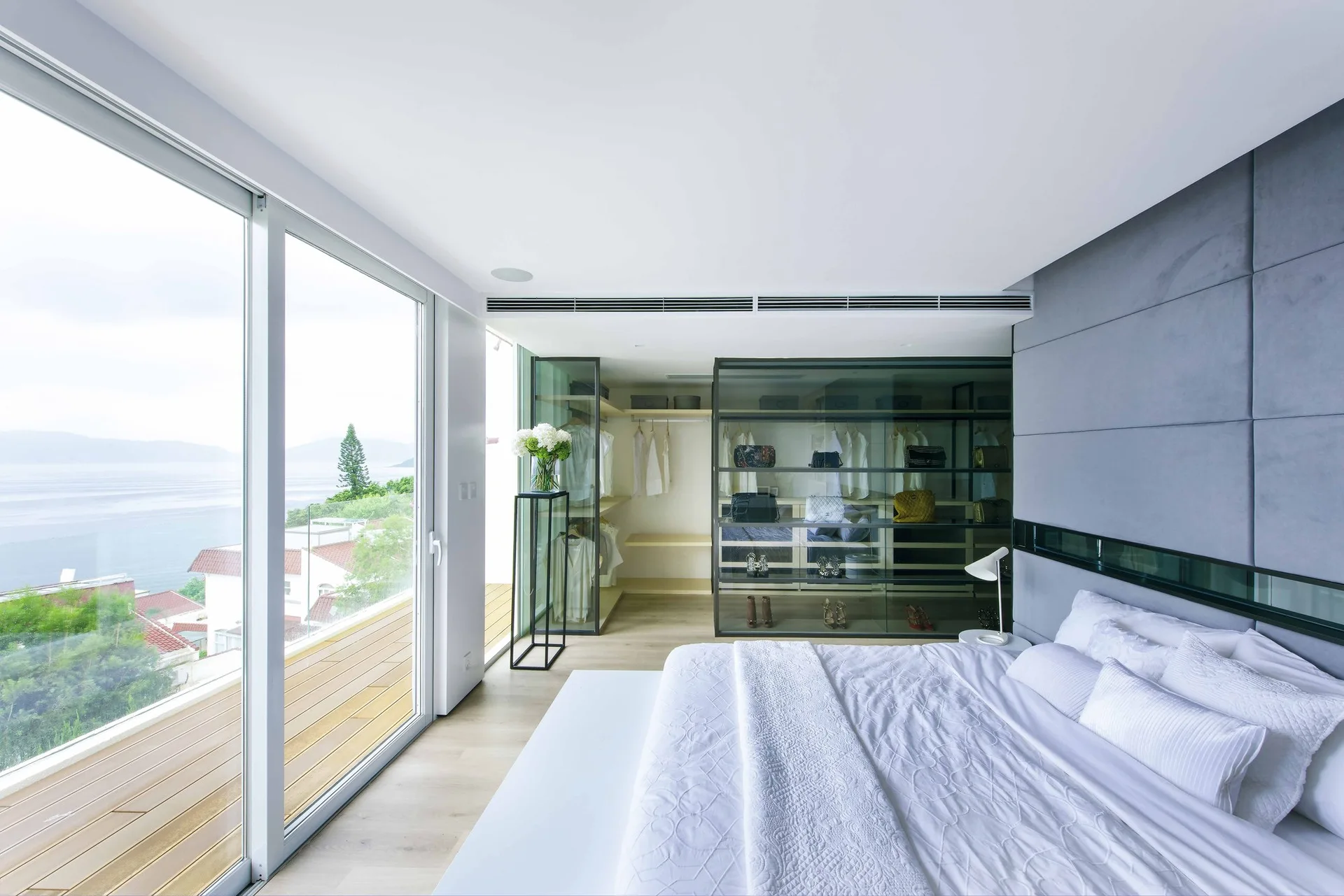 A minimalist bedroom featuring a bed with white bedding, a grey upholstered wall panel, a glass-front wardrobe, and sliding doors to a scenic balcony.