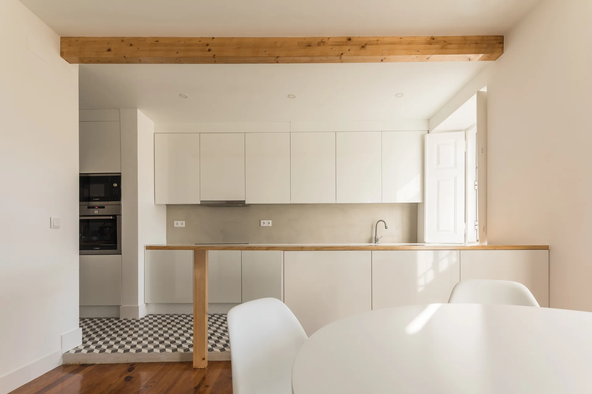 A minimalist white kitchen features flat-front cabinets, a light wood countertop, an exposed ceiling beam, and black-and-white geometric floor tiles.