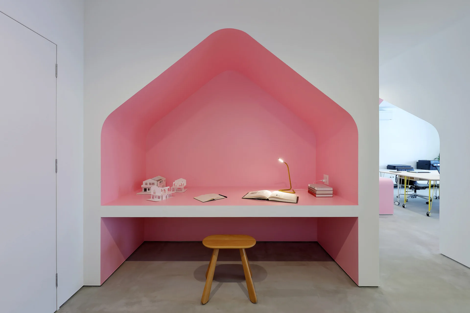 A built-in pink desk alcove shaped like a house features a small wooden stool, a task lamp, and white walls.