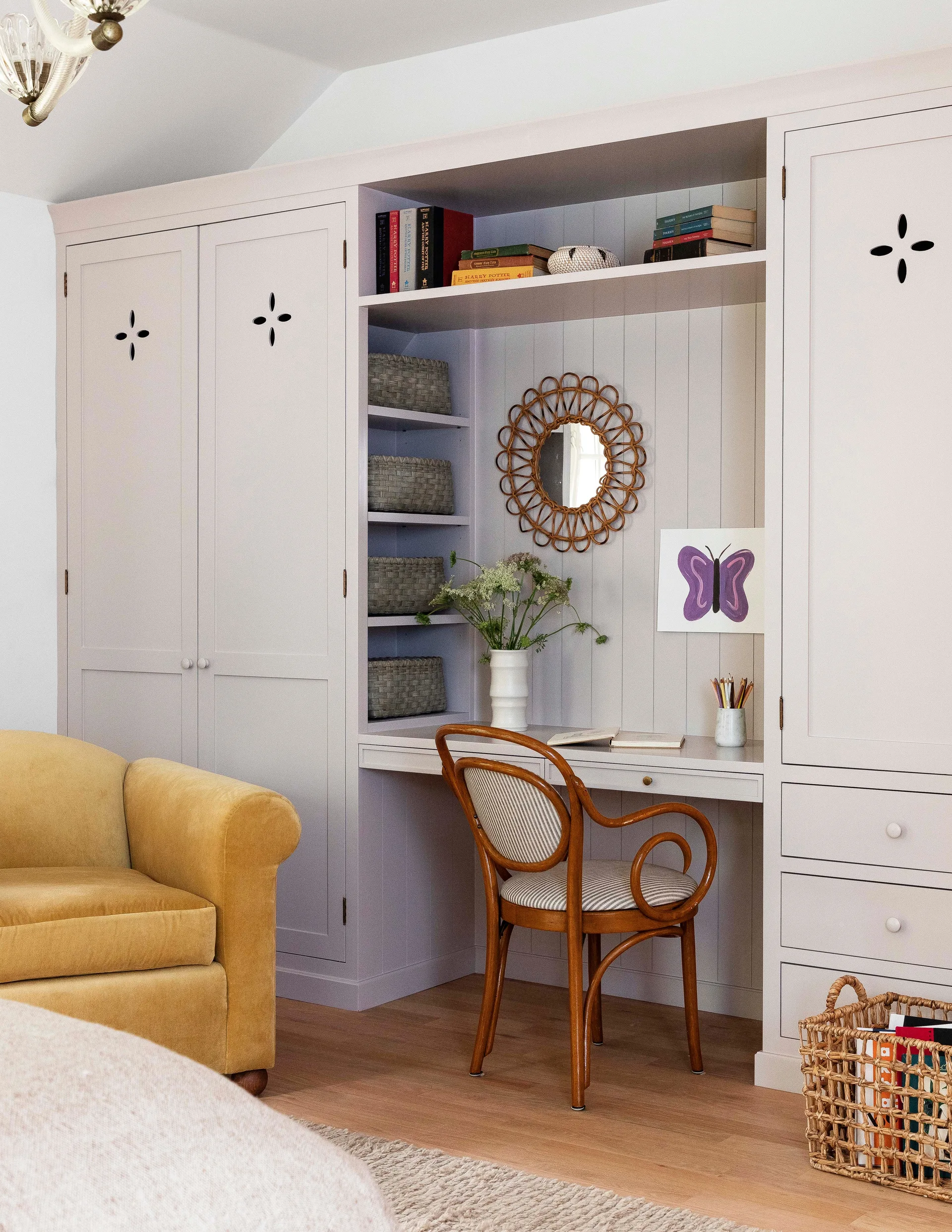 Lilac built-in bedroom cabinets frame a small desk with a bentwood chair, a rattan mirror, and a mustard yellow armchair.