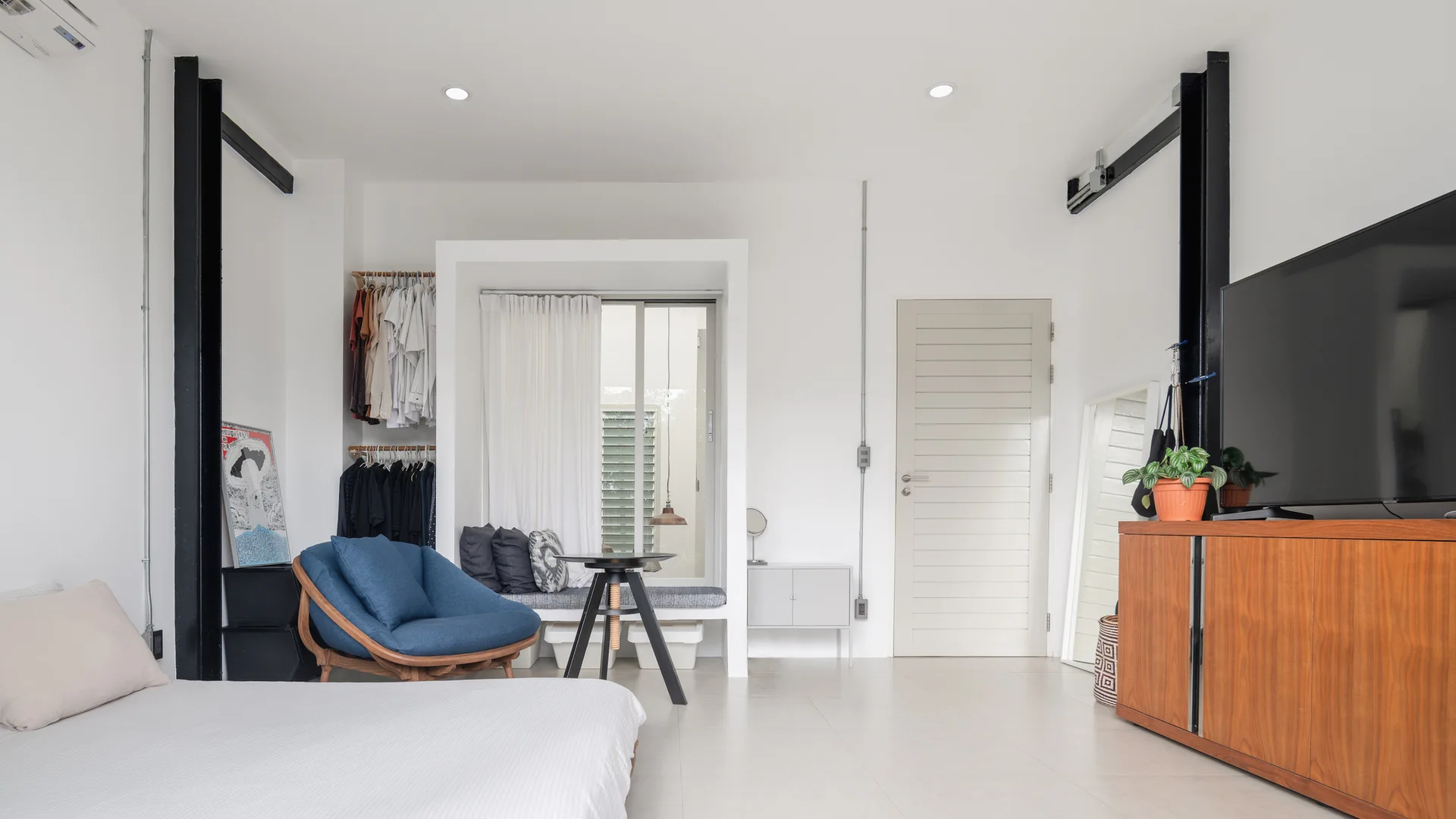 A minimalist bedroom featuring a built-in window bench, a blue armchair, an open clothing rack, and a wood media console.