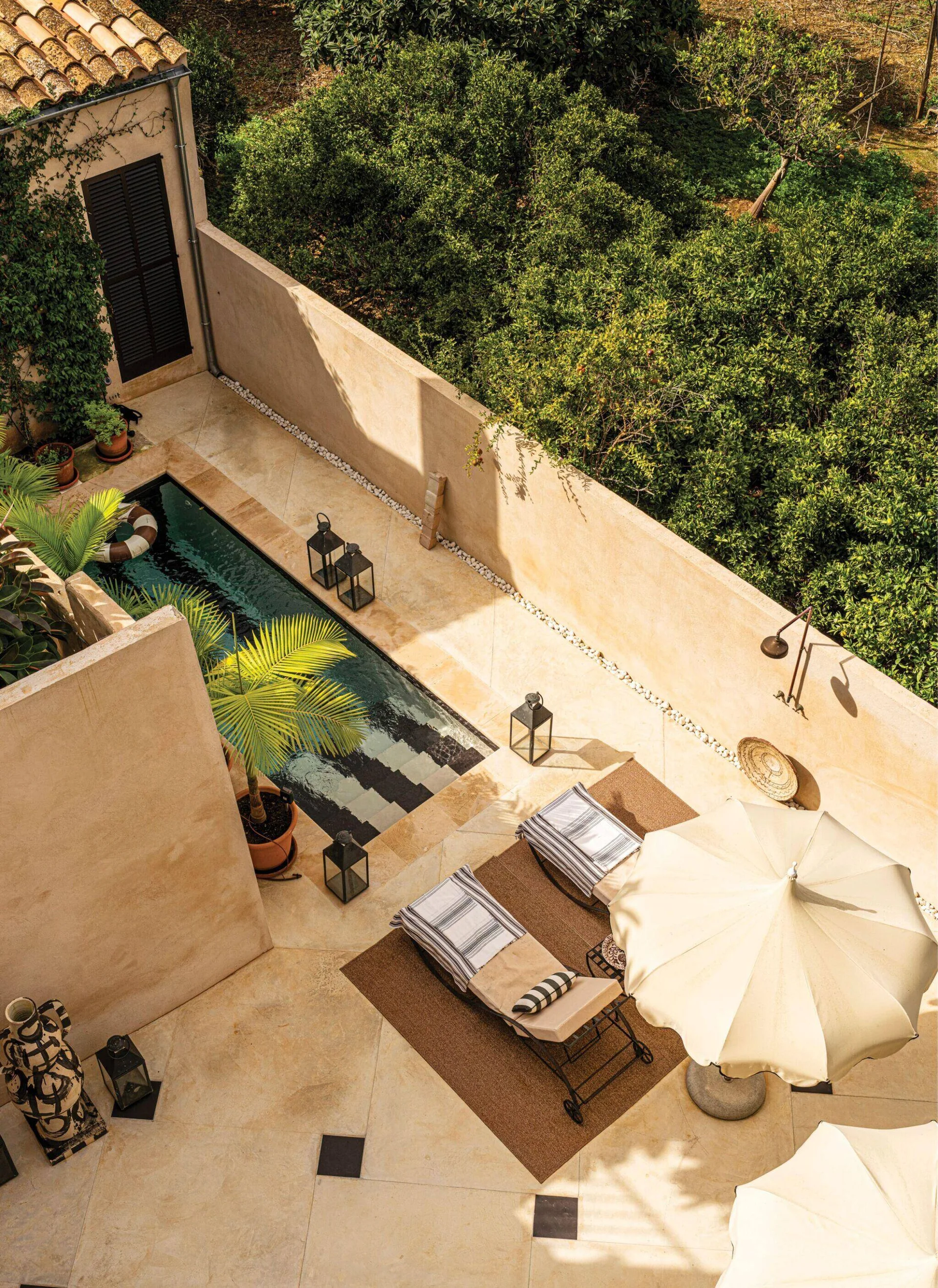 An overhead view of a sunny courtyard featuring a narrow lap pool, beige stone tiles, stucco walls, and two sun loungers under a cream umbrella.