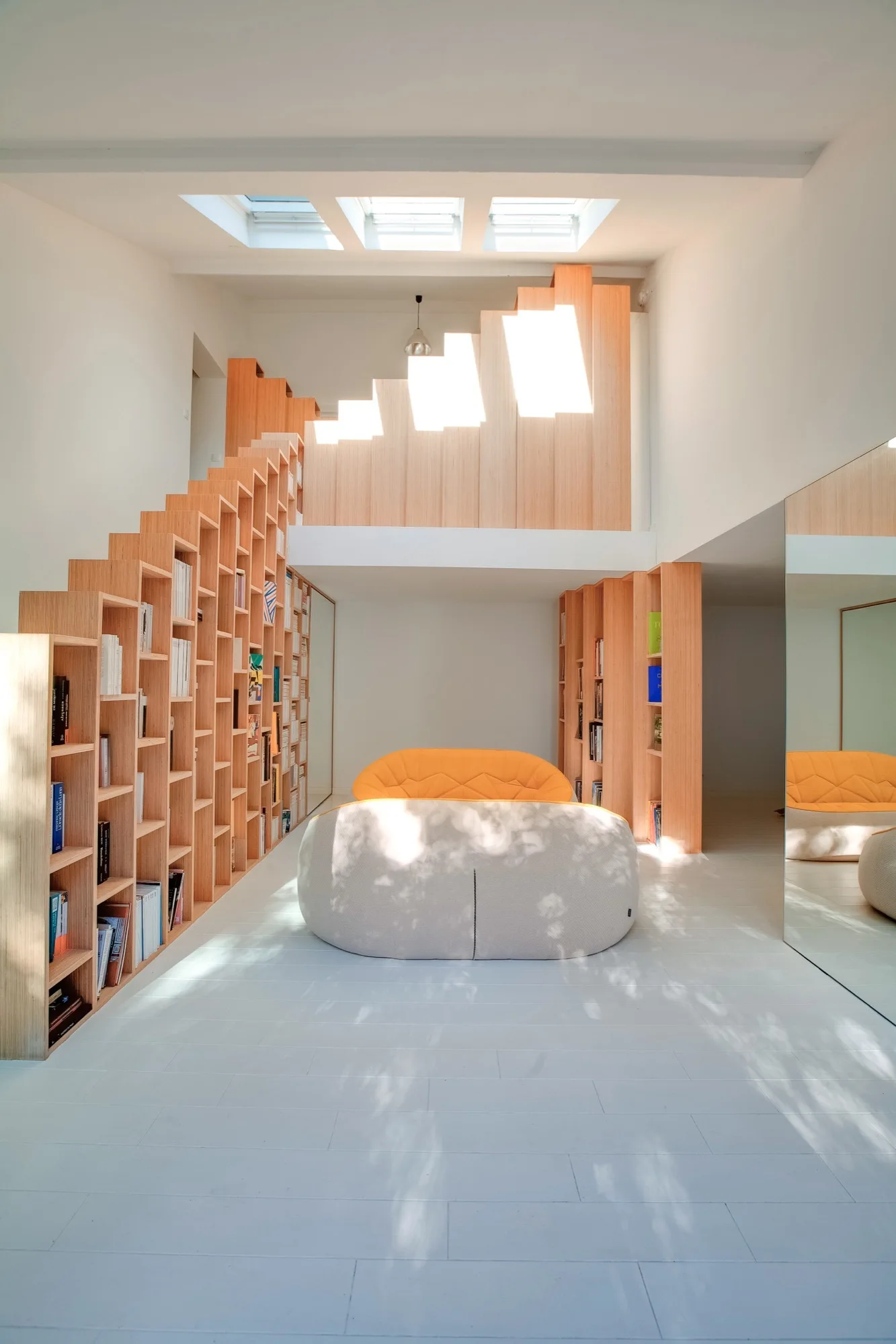 A modern white living room featuring a light wood staircase with built-in bookshelves, a curved cream and orange sofa, and skylights.