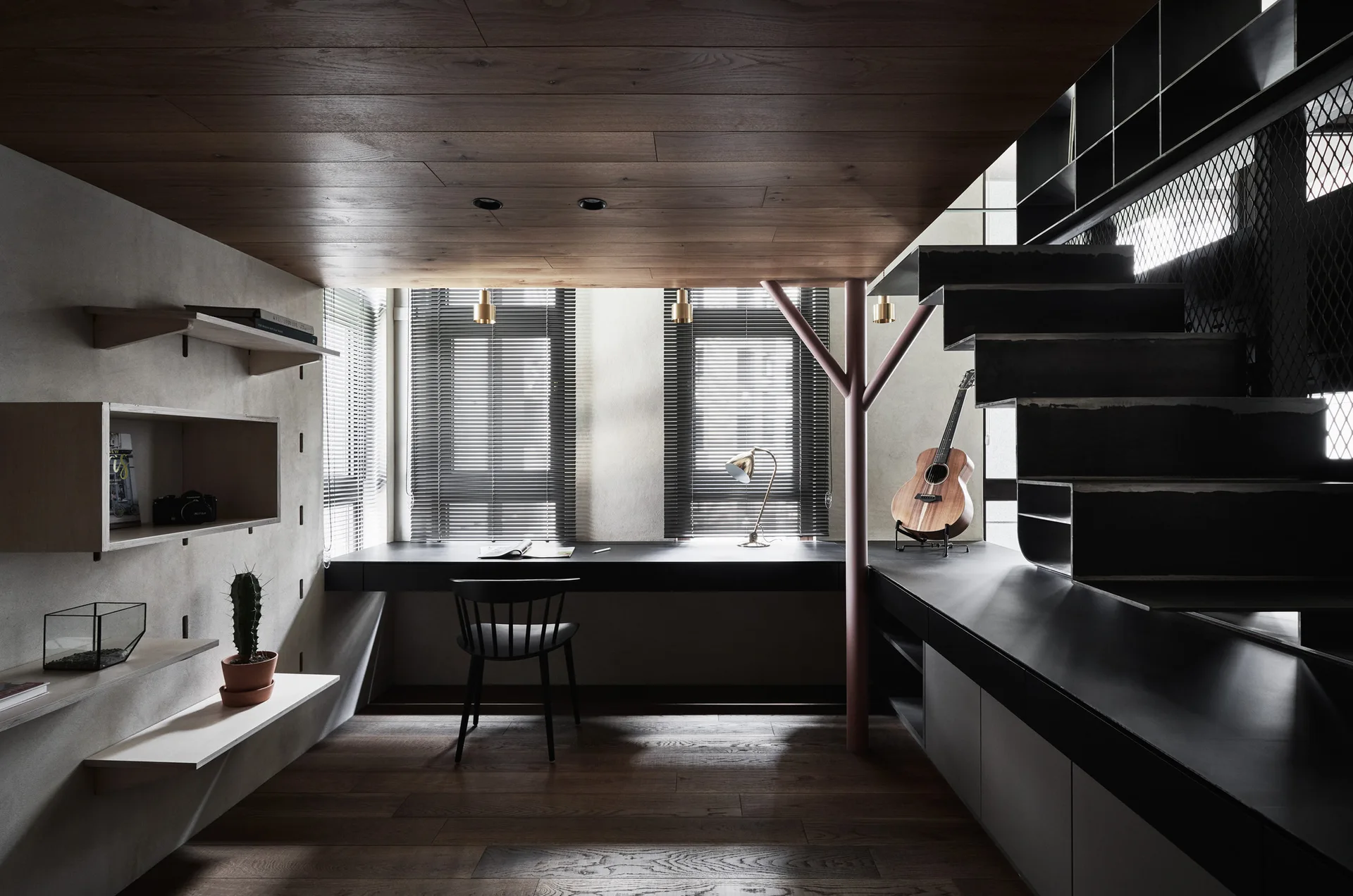 A home office features a dark integrated desk under windows, a wood paneled floor and ceiling, floating shelves, and a metal staircase.