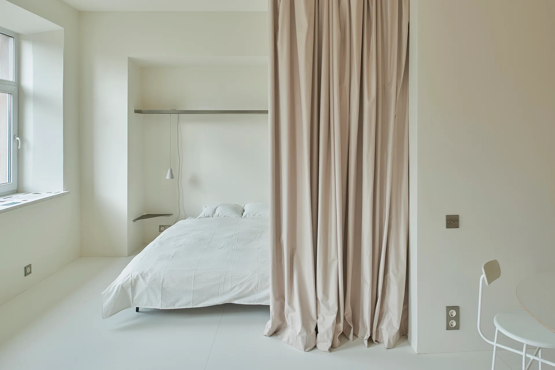 A minimalist white bedroom features a bed tucked into a recessed alcove with a long beige fabric curtain acting as a partition.