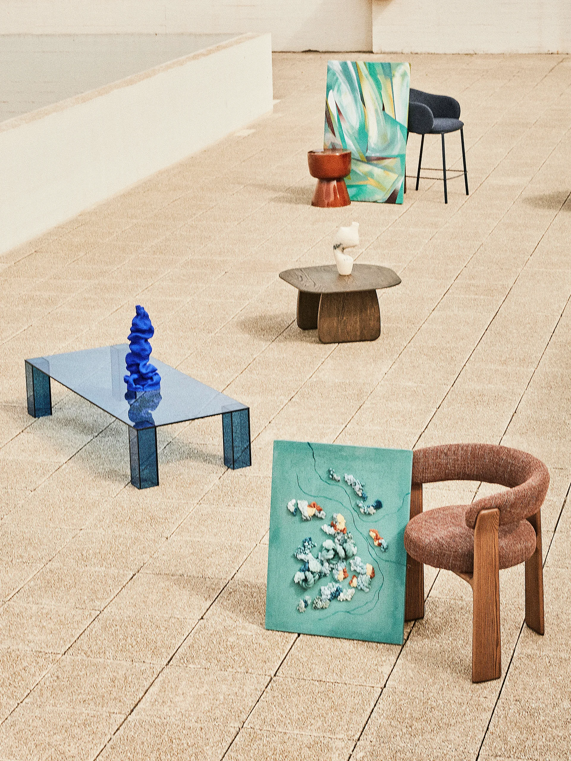 Modern furniture pieces including a blue glass coffee table and a brown upholstered armchair are displayed on a sandy-colored tiled outdoor floor.
