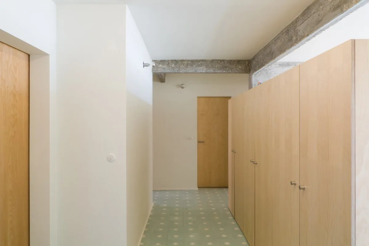 A modern residential hallway features light wood built-in cabinets, a patterned green tile floor, and an exposed concrete ceiling beam.
