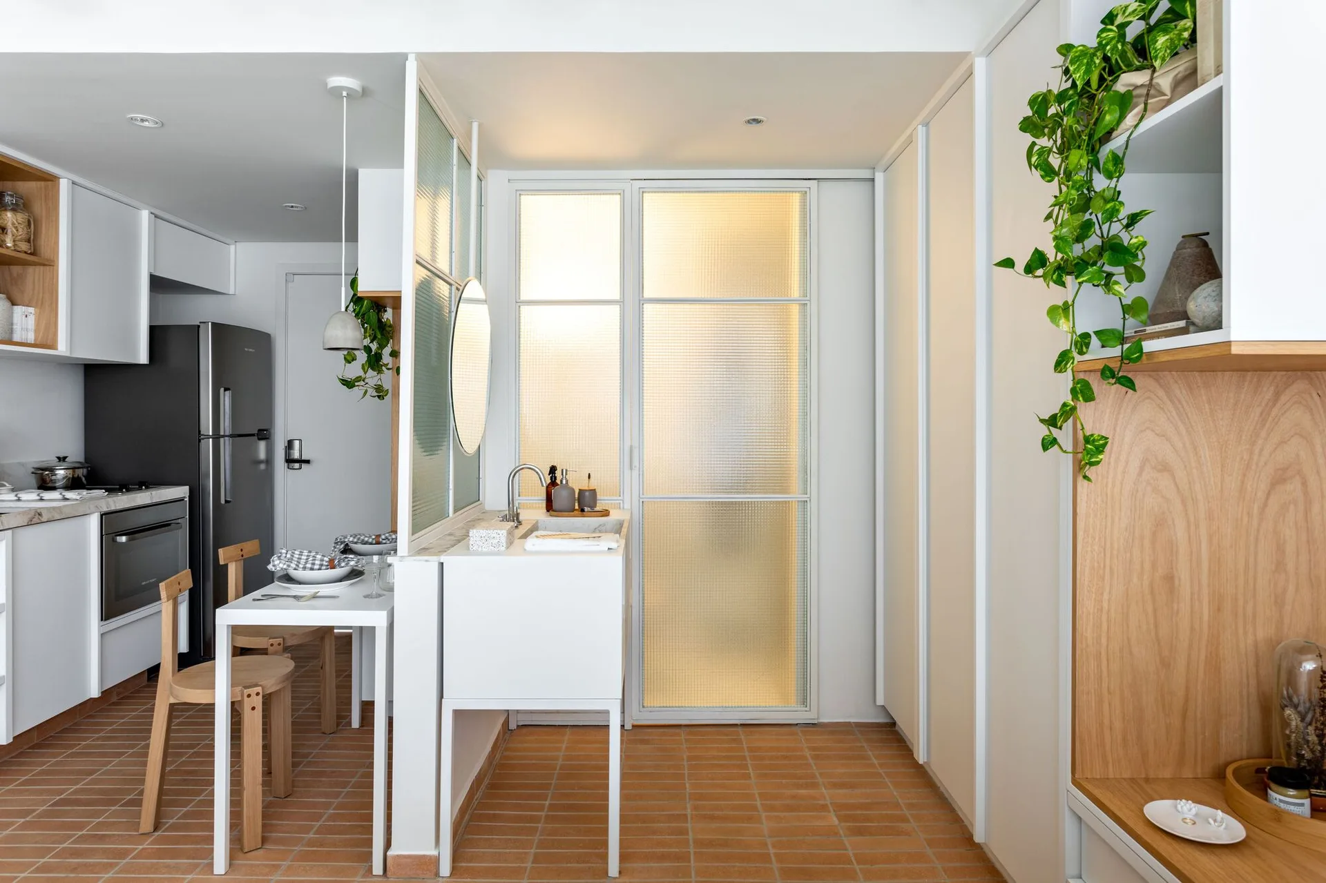 A compact kitchen and dining area featuring terracotta floors, white cabinetry with light wood accents, and a translucent textured glass partition.