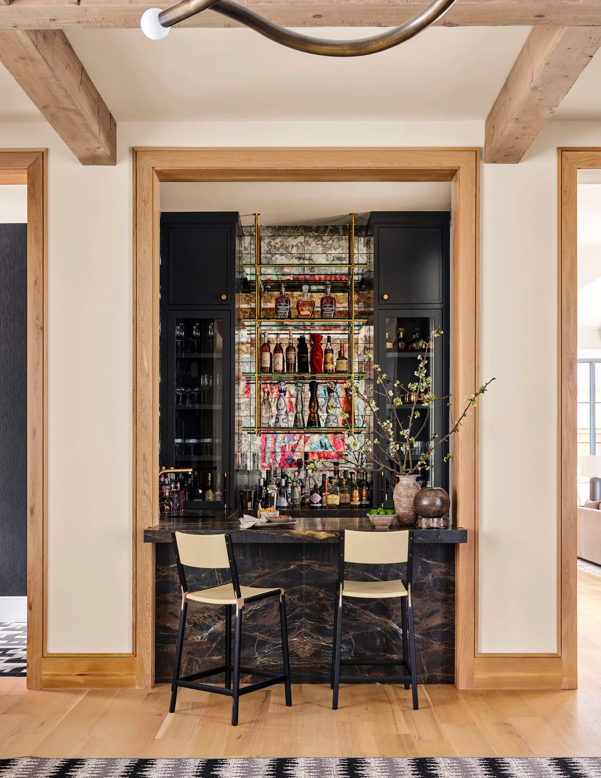 A built-in home bar features black cabinetry, an antique mirror backsplash with brass shelves, a dark stone counter, and two bar stools.