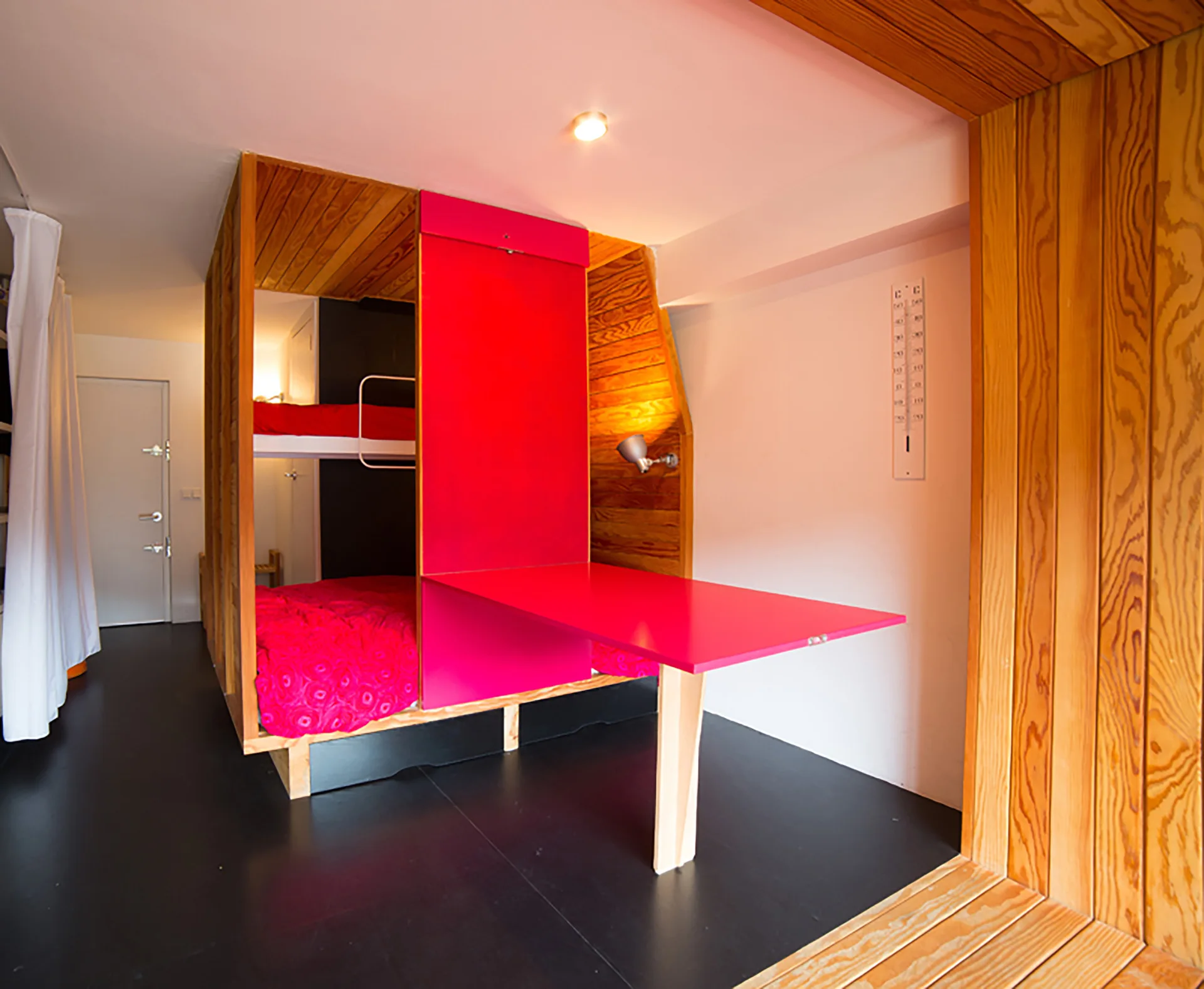 A compact bedroom features a built-in wooden bunk bed structure integrated with a bright pink folding desk and dark flooring.
