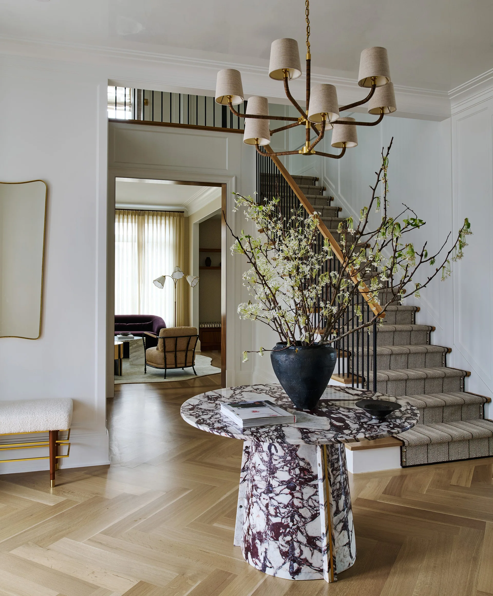 A bright entryway features a round purple-veined marble table, a staircase with a patterned runner, and herringbone wood flooring.