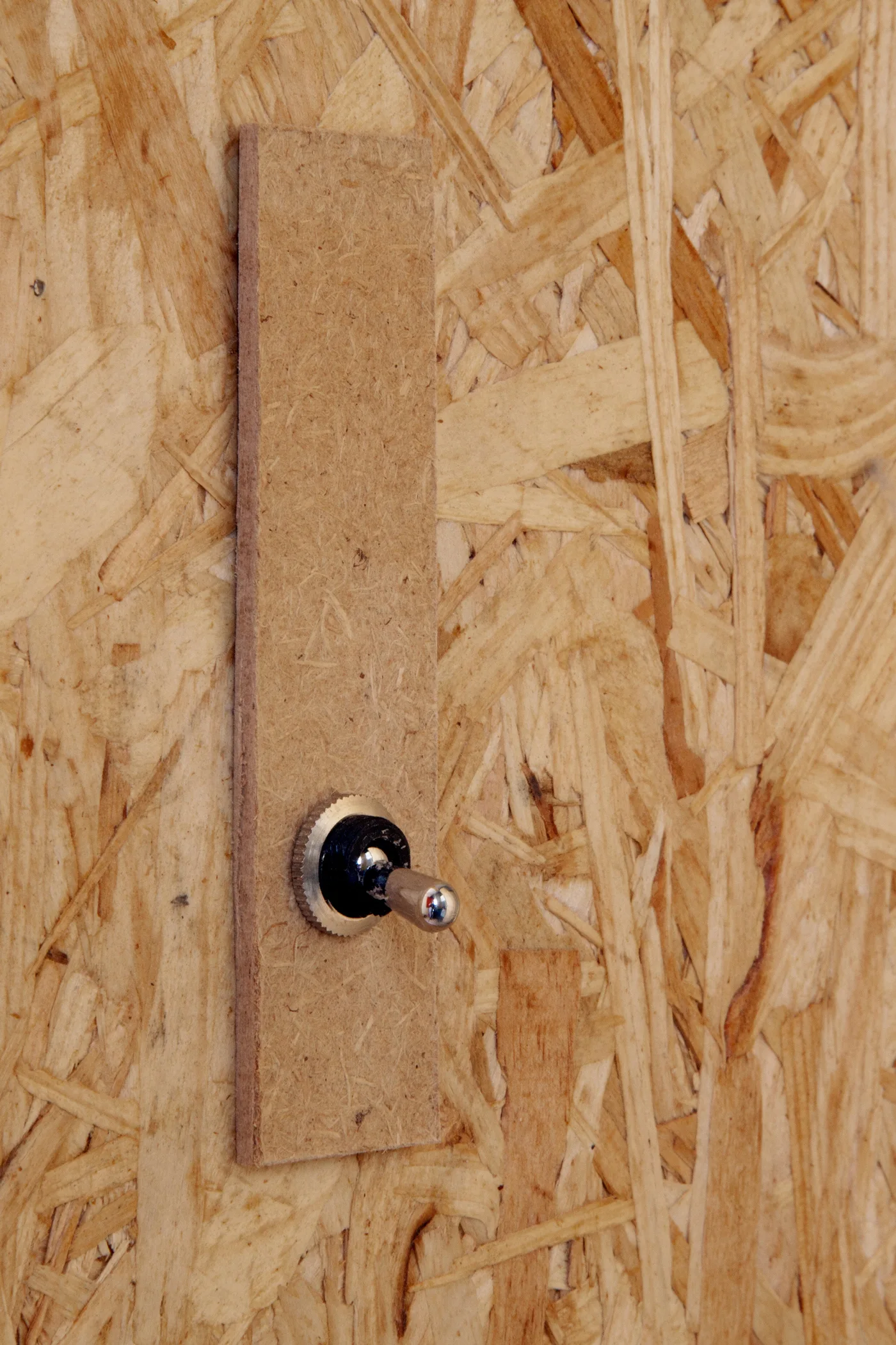 A metallic toggle switch mounted on a narrow wood-particle backing plate against an oriented strand board wall.
