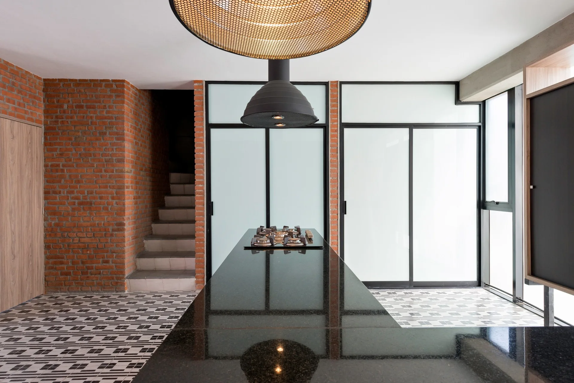 A modern industrial kitchen featuring a polished black stone island with a cooktop, geometric floor tiles, and an exposed red brick wall.