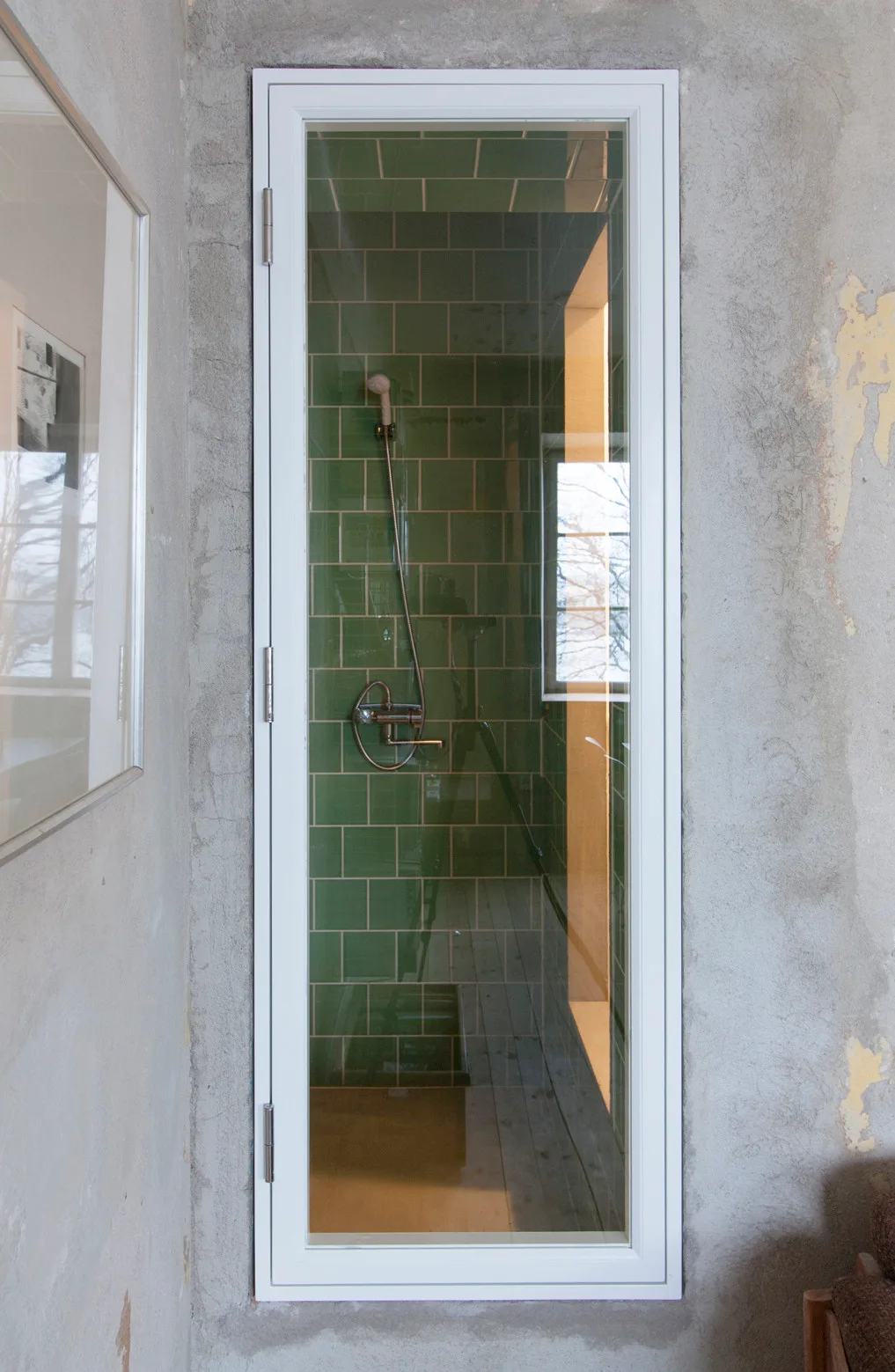 A narrow glass shower door with a white frame is set into a raw concrete wall, showing glossy green tiles inside.
