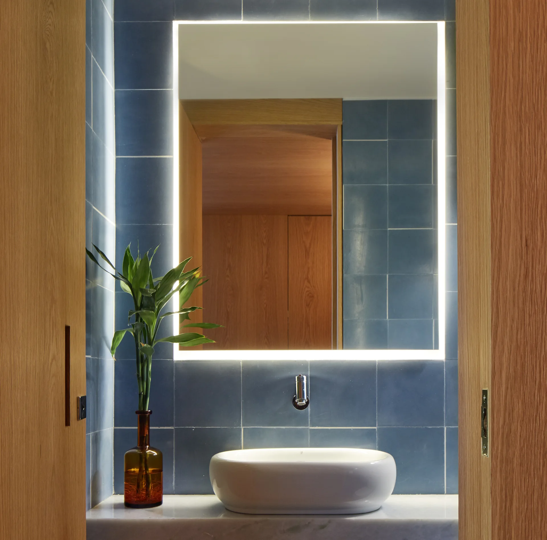 A white vessel sink rests on a marble vanity in front of a blue tiled wall and a rectangular backlit mirror, framed by natural wood doors.