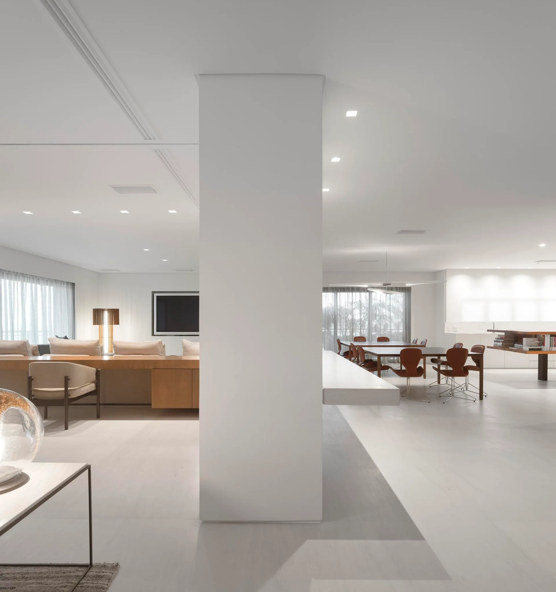 A large minimalist living and dining space features a central white structural column, neutral upholstered sofa, and wood dining set.