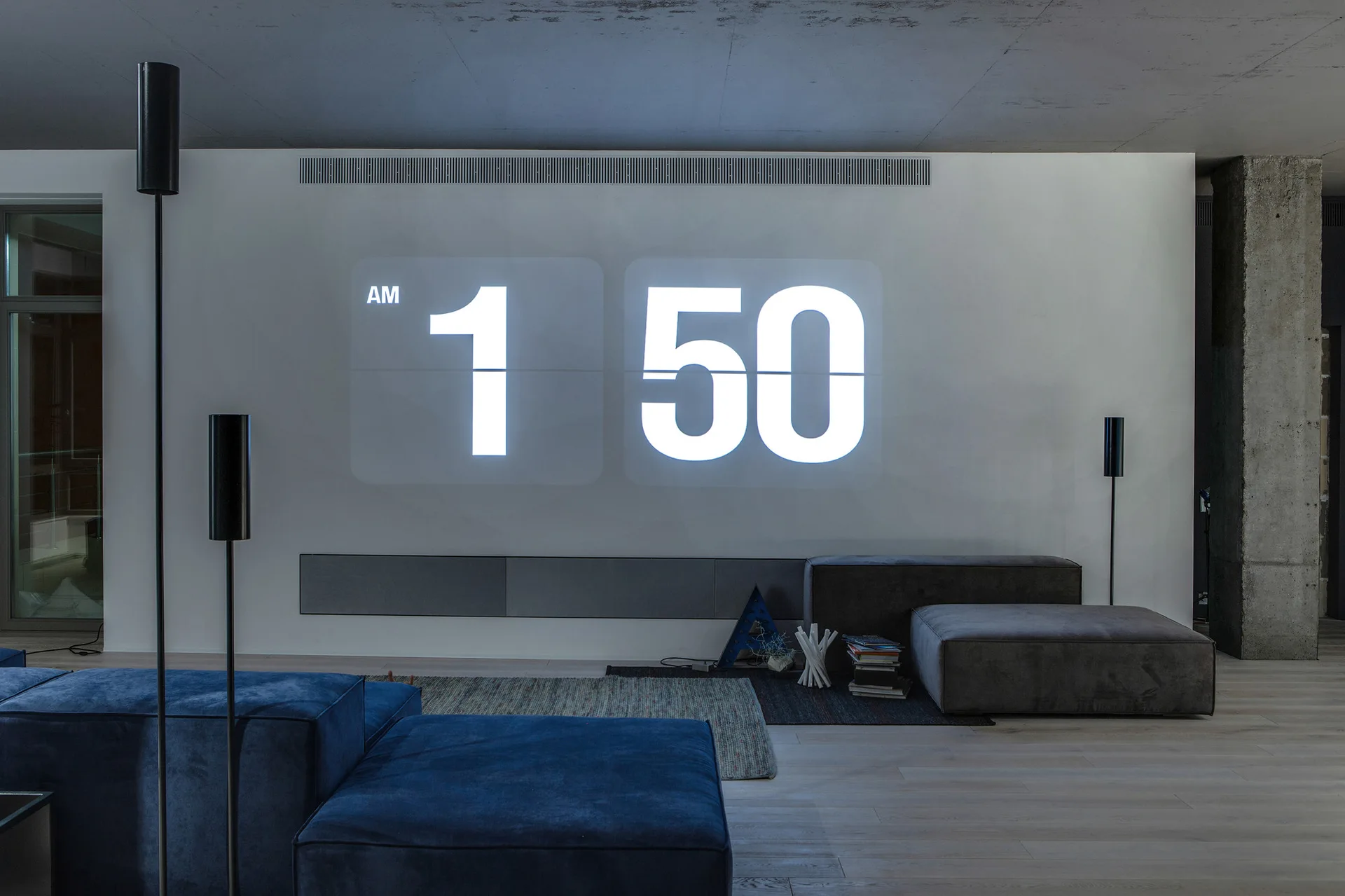 A modern living room features a blue modular sofa, grey low-profile seating, and a large digital flip-clock projected onto a white wall.