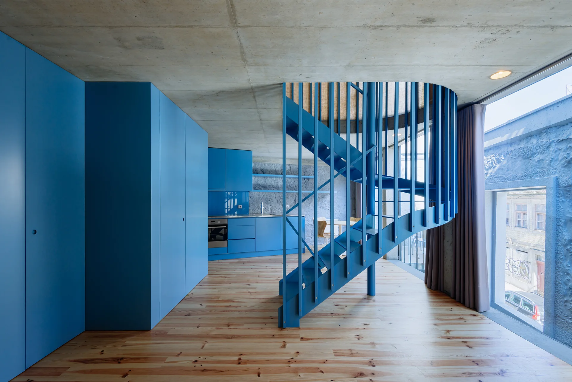 A modern loft interior with light wood flooring, an exposed concrete ceiling, and a prominent blue spiral staircase next to matching blue cabinets.