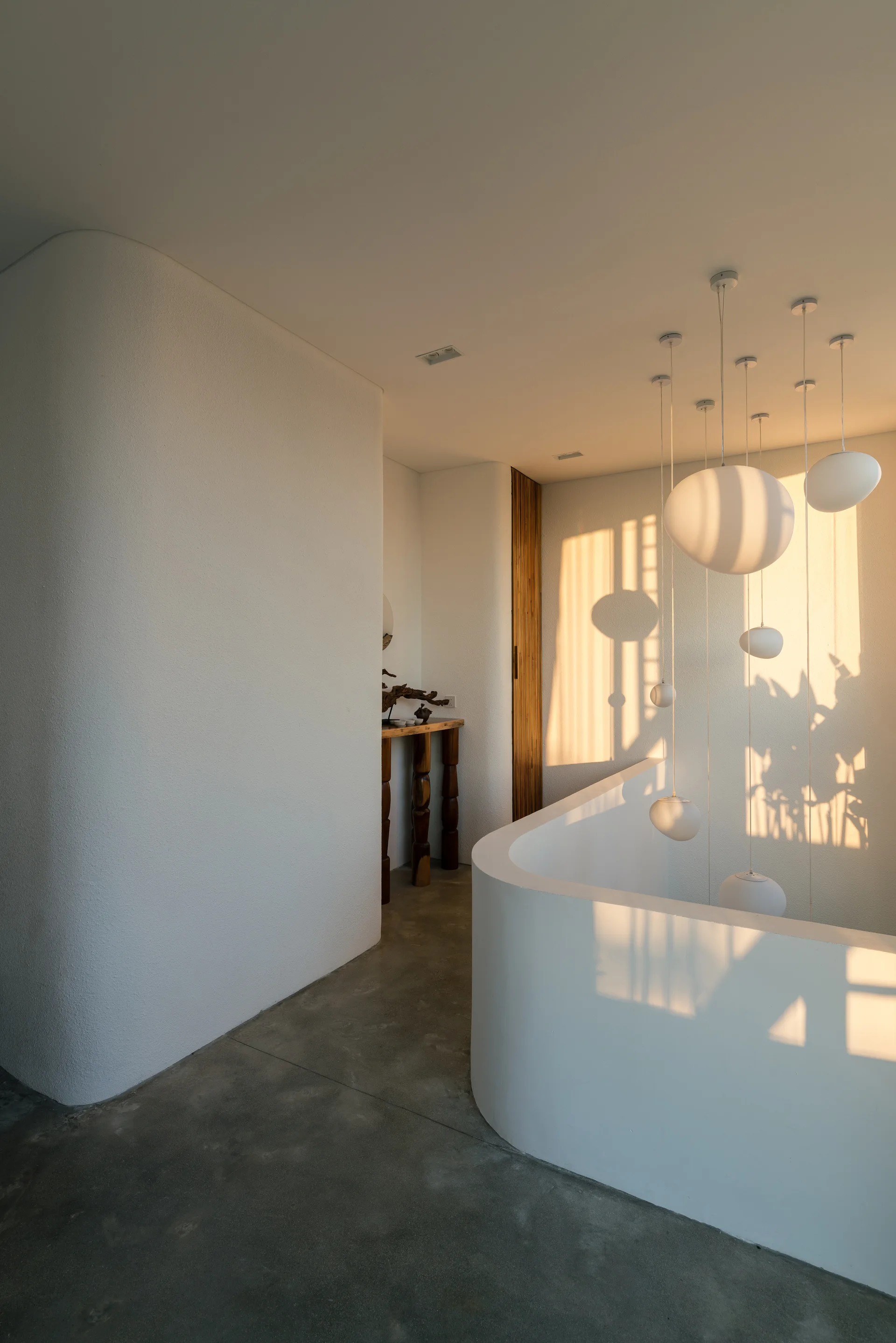 A minimalist staircase landing features curved plaster walls, concrete floors, and a cluster of white globe pendant lights catching warm sunset light.