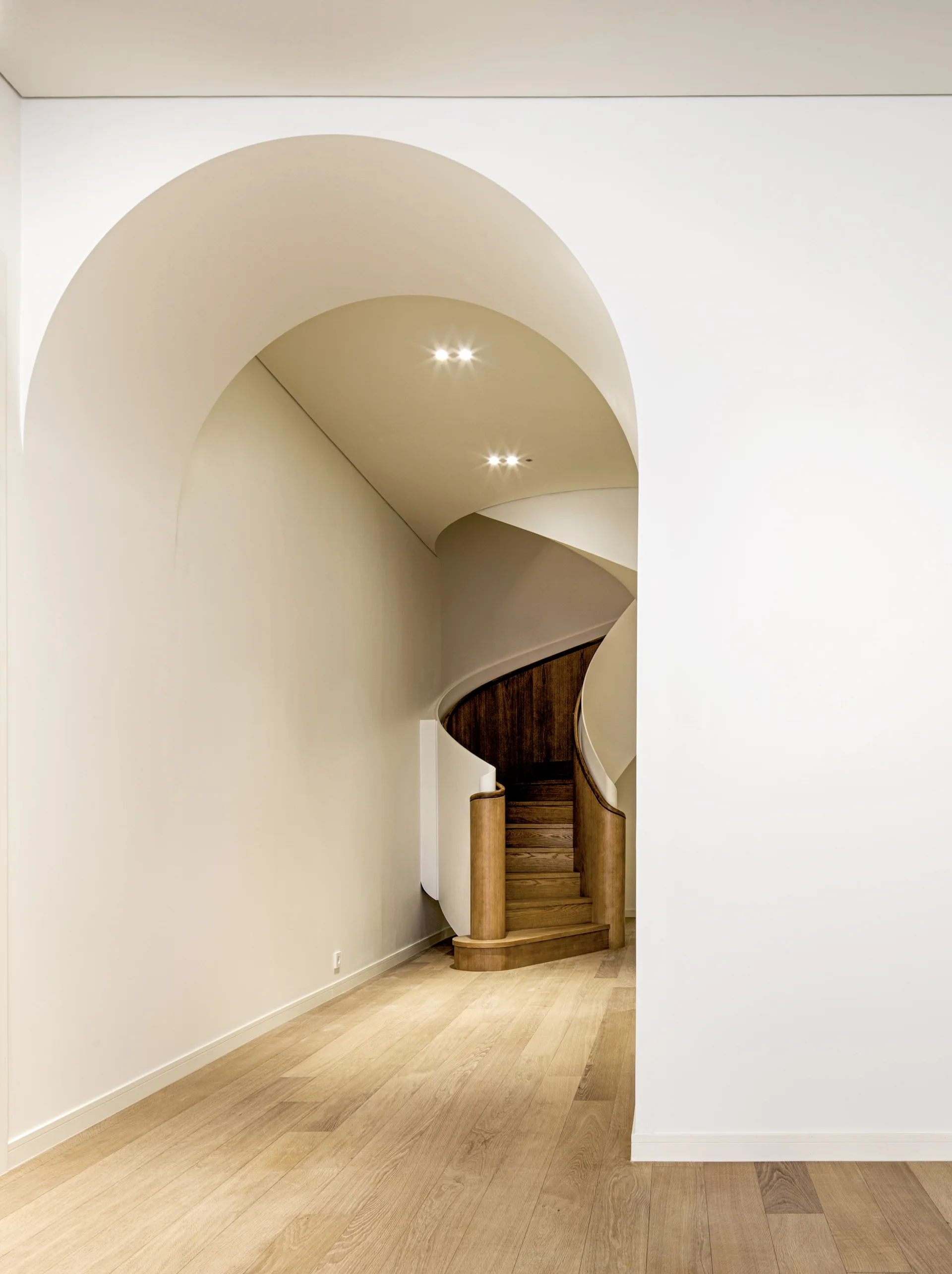 A white arched entryway frames a modern curved wooden staircase with a smooth white balustrade under recessed lighting.