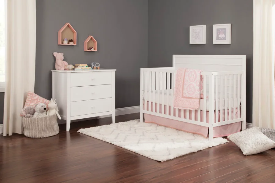 A white wooden crib and matching dresser stand against dark grey walls in a nursery decorated with pink textiles and a textured area rug.