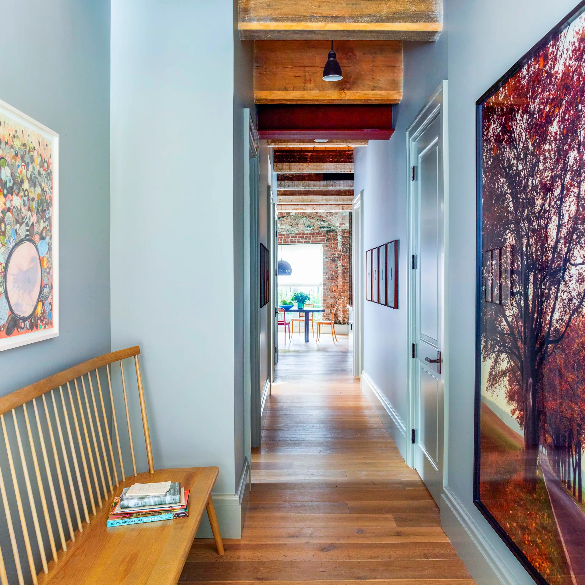 A long blue-gray hallway features exposed wood ceiling beams, a light wood spindle bench, and hardwood flooring leading to a brick-walled room.