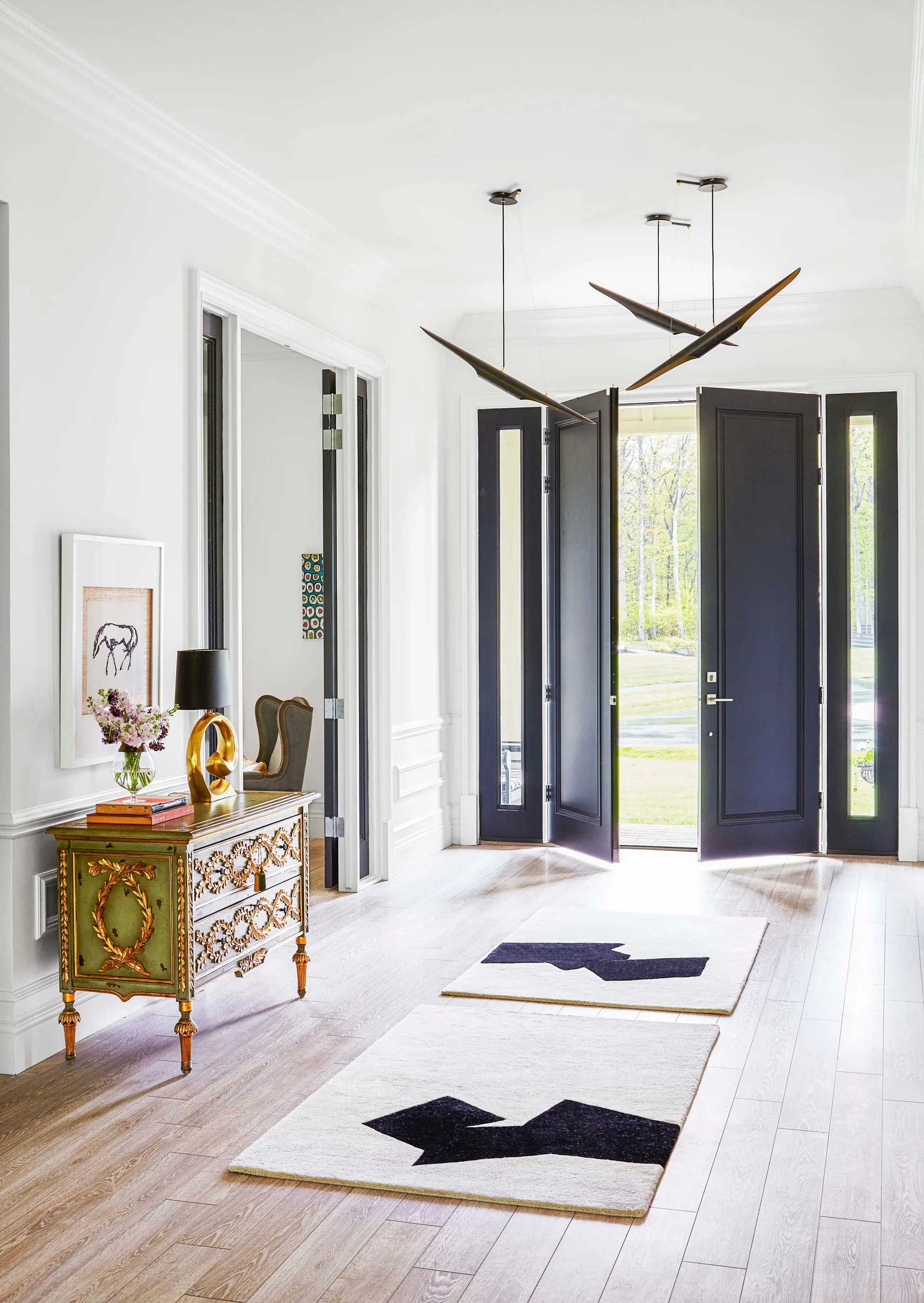 A bright entryway features charcoal double doors, light wood flooring, two geometric rugs, and an ornate green and gold chest of drawers.