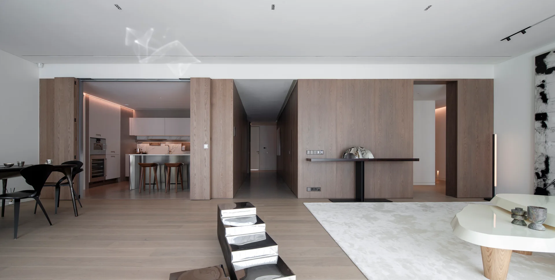 A modern minimalist living room features warm wood paneled walls, light wood floors, and a view into an adjacent kitchen.