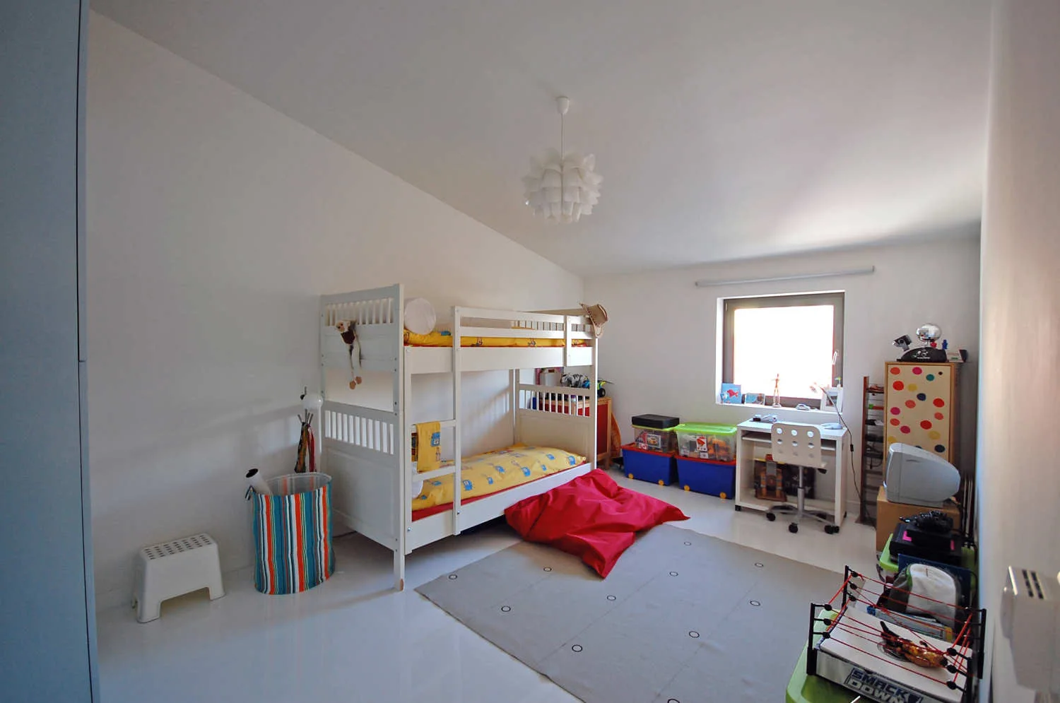 A bright children's bedroom features a white wooden bunk bed, a desk under a window, a red bean bag, and a grey rug on a glossy white floor.