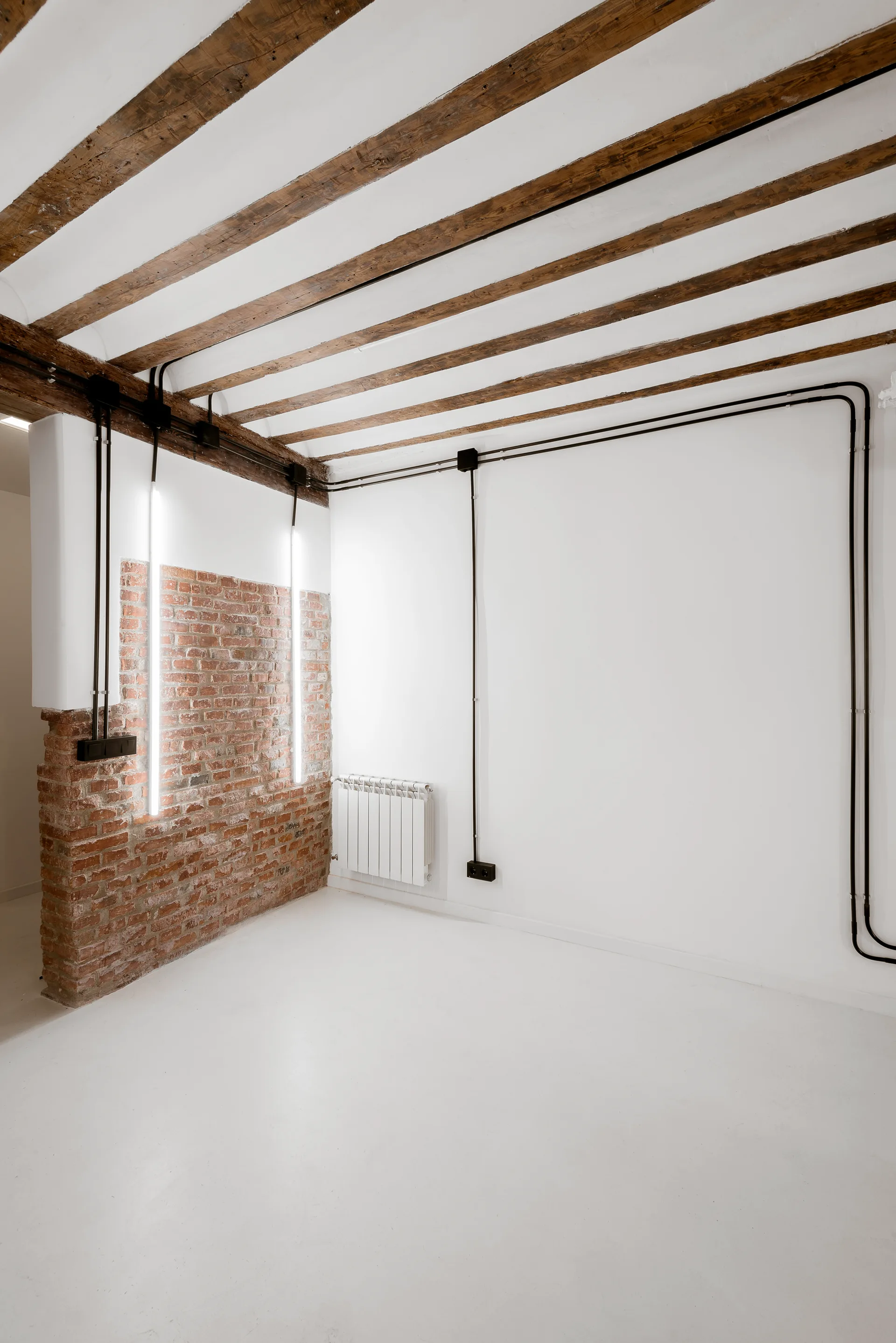 An empty industrial style room with dark wood ceiling beams, white walls, black electrical conduits, and a red brick wall segment.