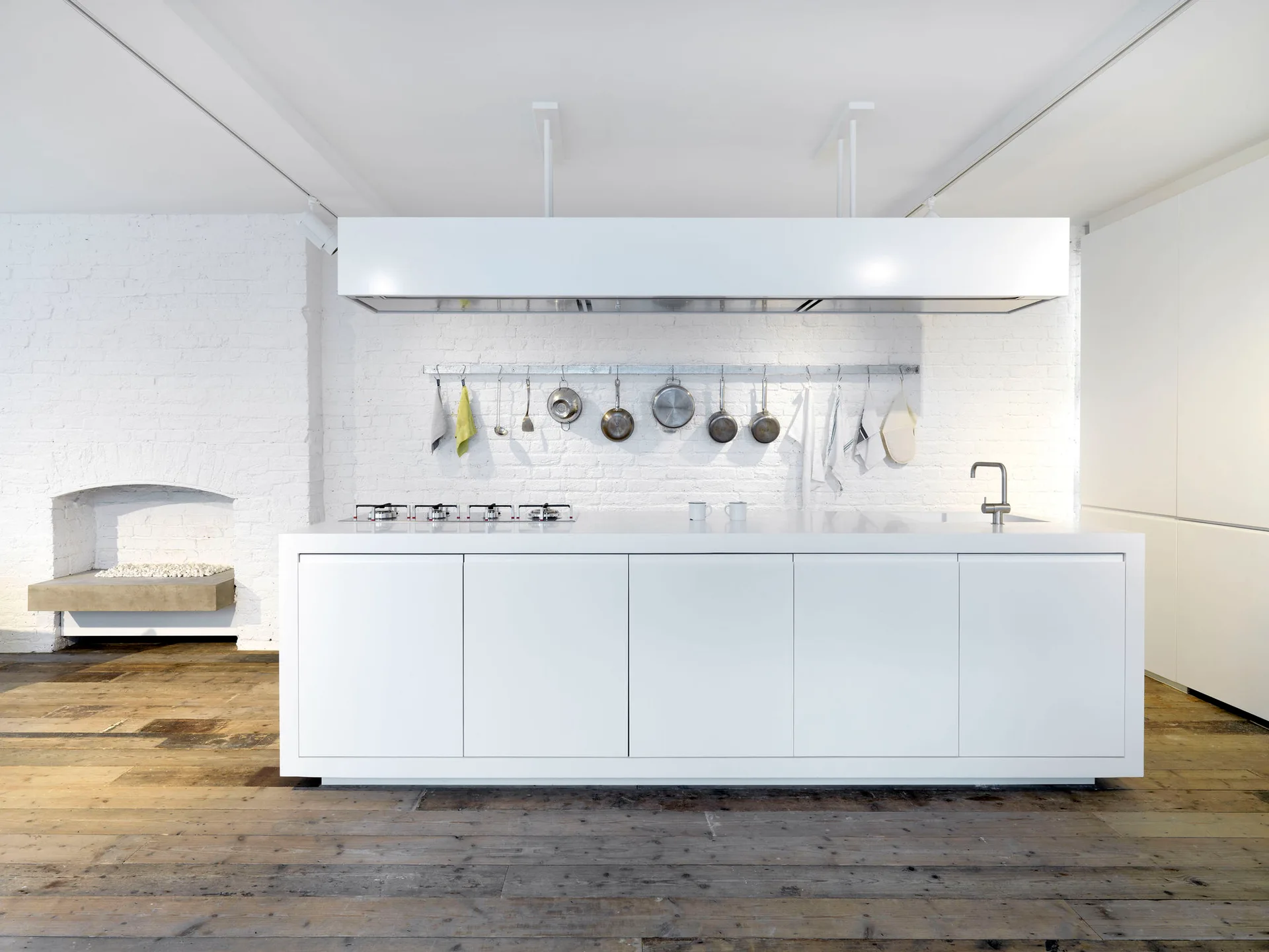 A minimalist white kitchen island with an integrated cooktop stands on weathered wood flooring before a painted white brick wall.
