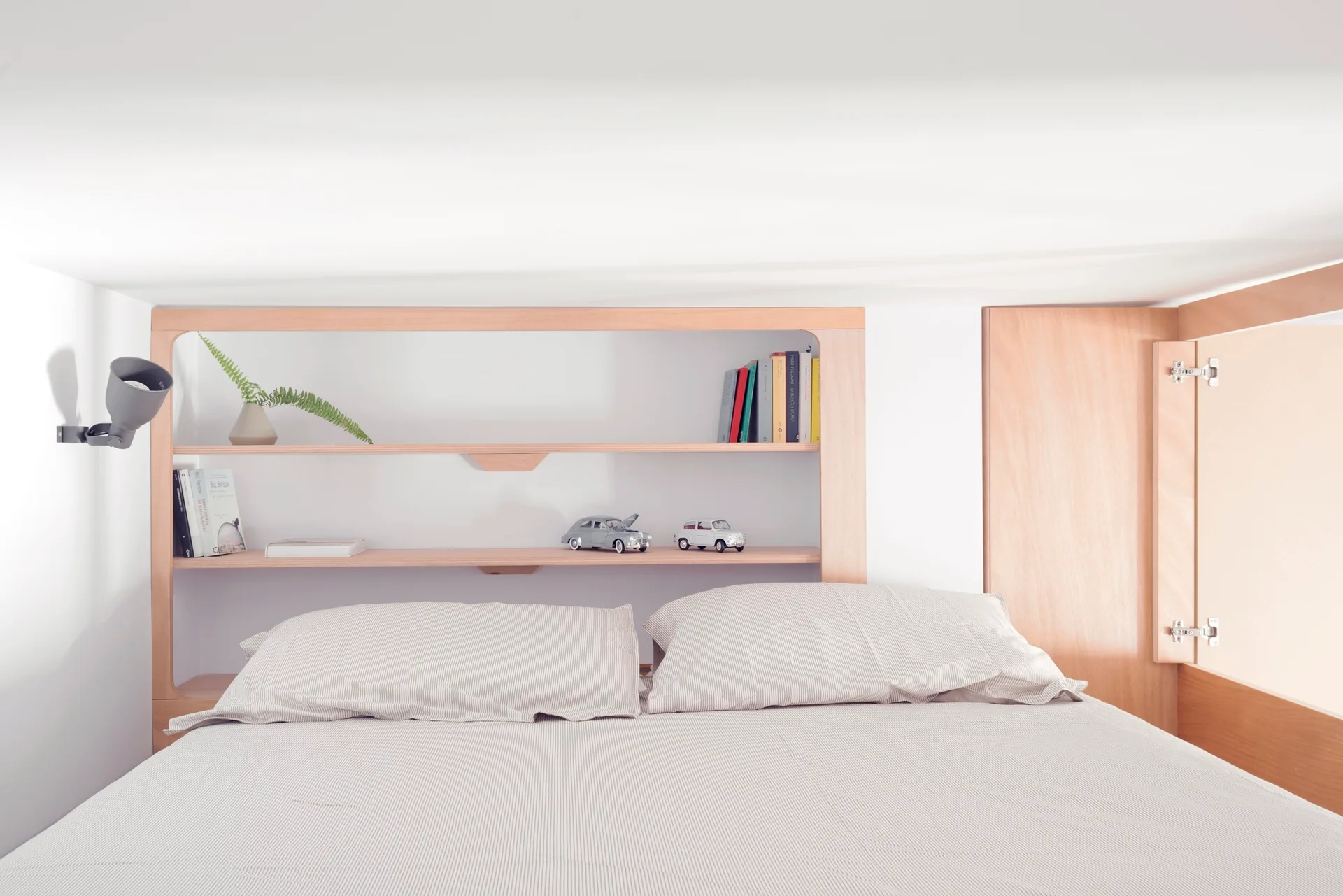 A bed with pinstripe linens sits in front of a custom light wood shelving headboard filled with books and small decor items.