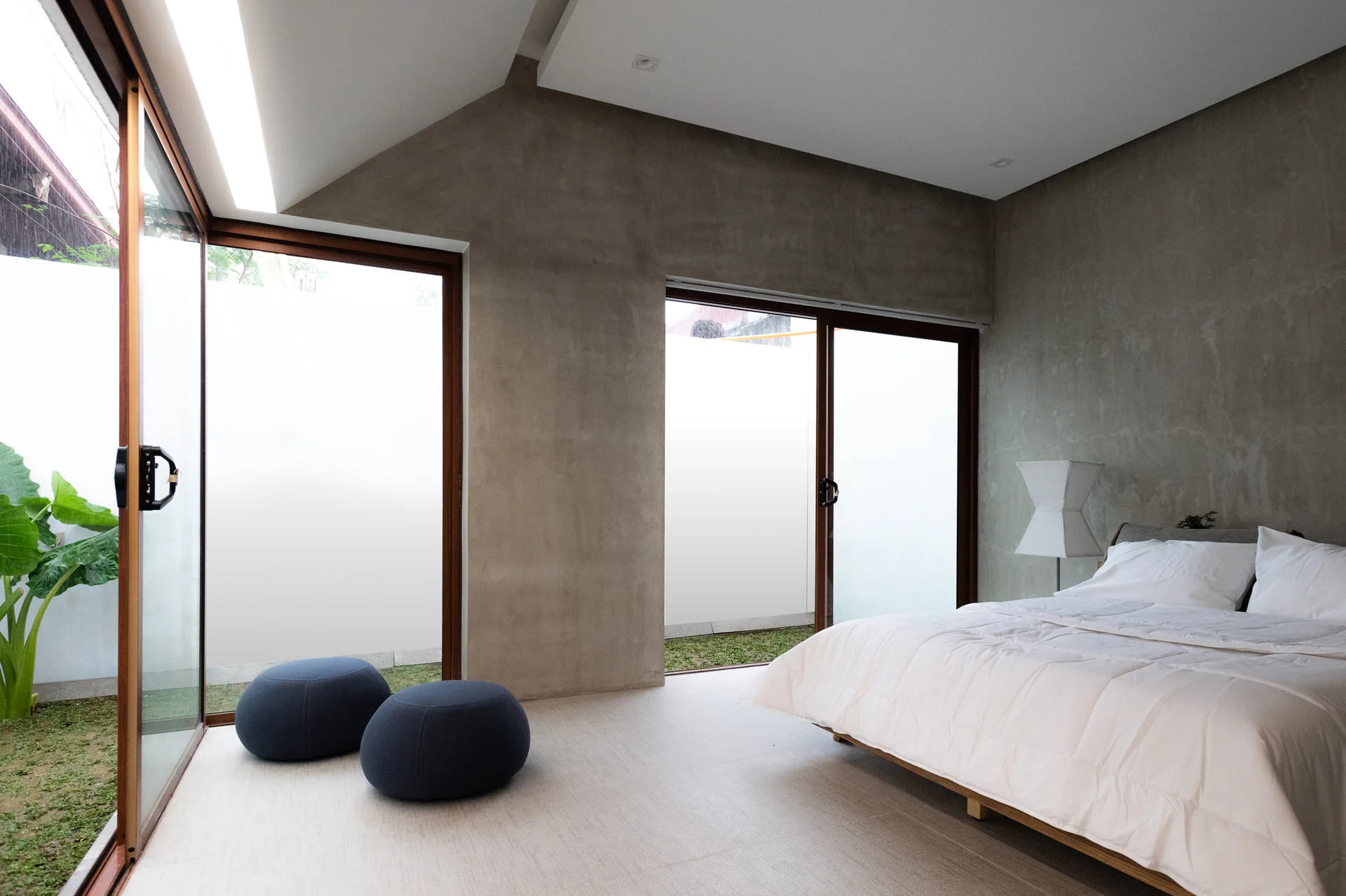 A minimalist bedroom featuring textured concrete walls, low blue floor poufs, a white bed, and sliding glass doors opening onto a garden.