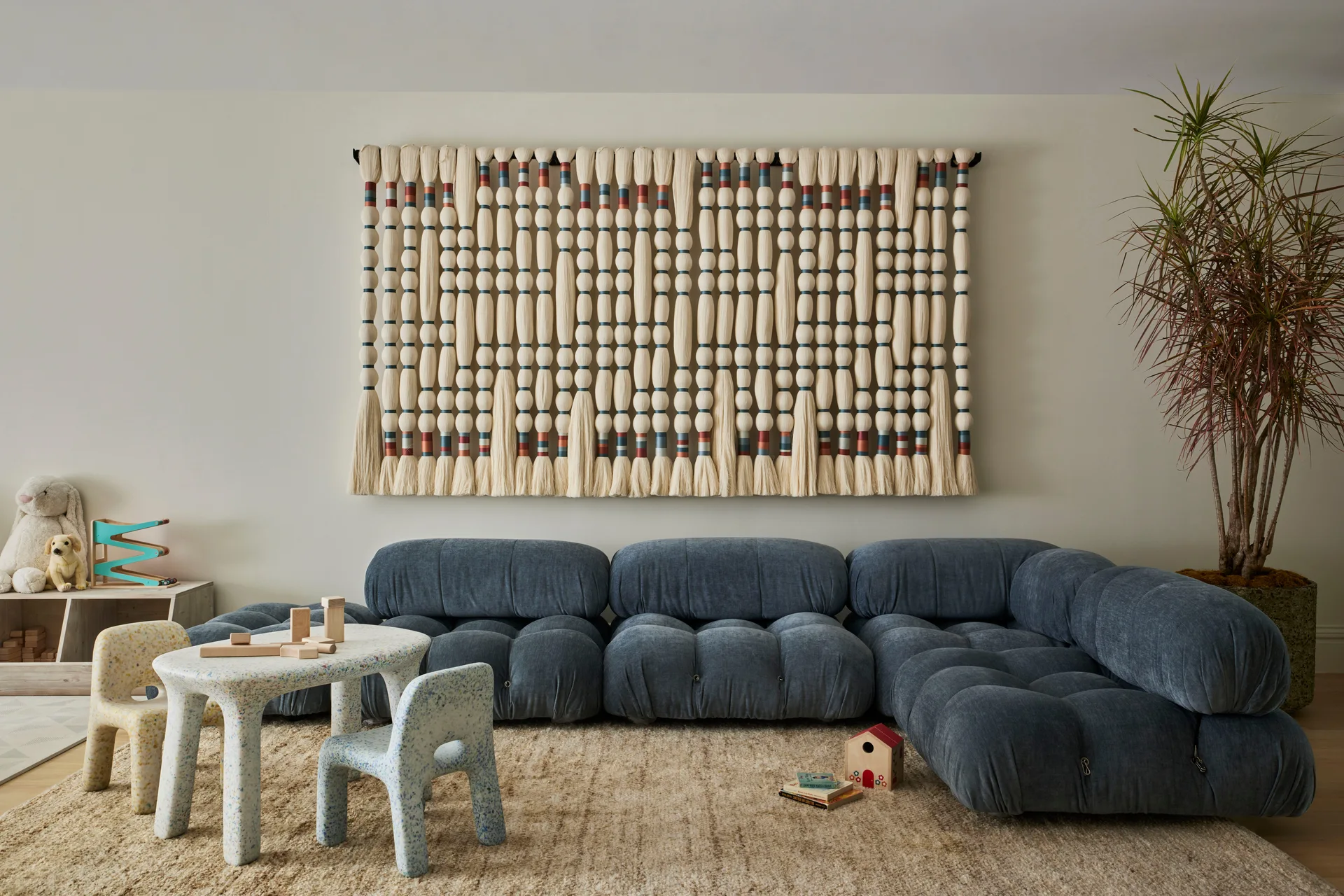 A children's playroom features a low blue modular sofa, a large knotted fiber wall hanging, and a speckled miniature table and chairs on a shag rug.