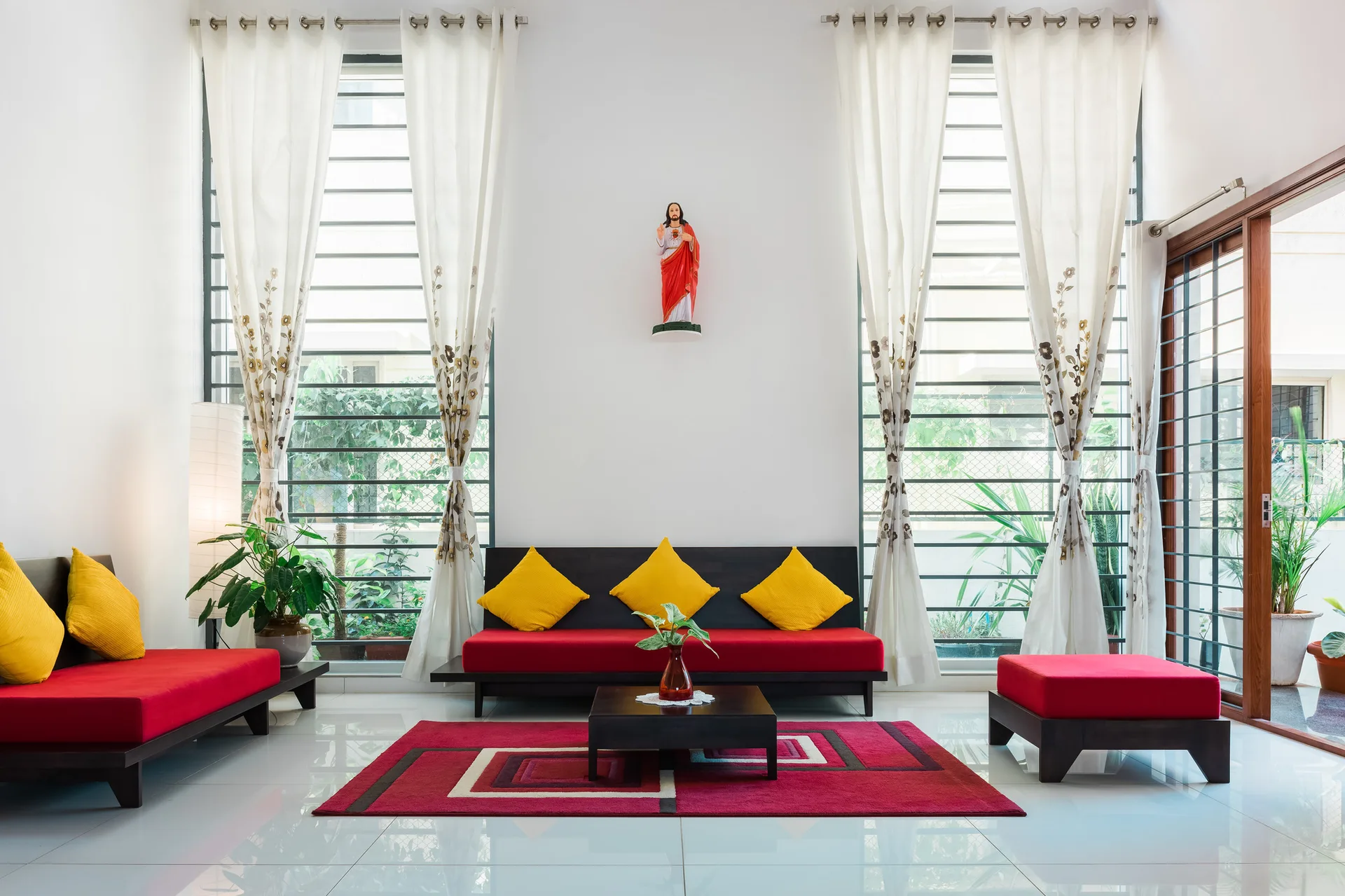 A symmetrical living room featuring red sofas with yellow pillows, a red geometric rug, and large windows with sheer white curtains.